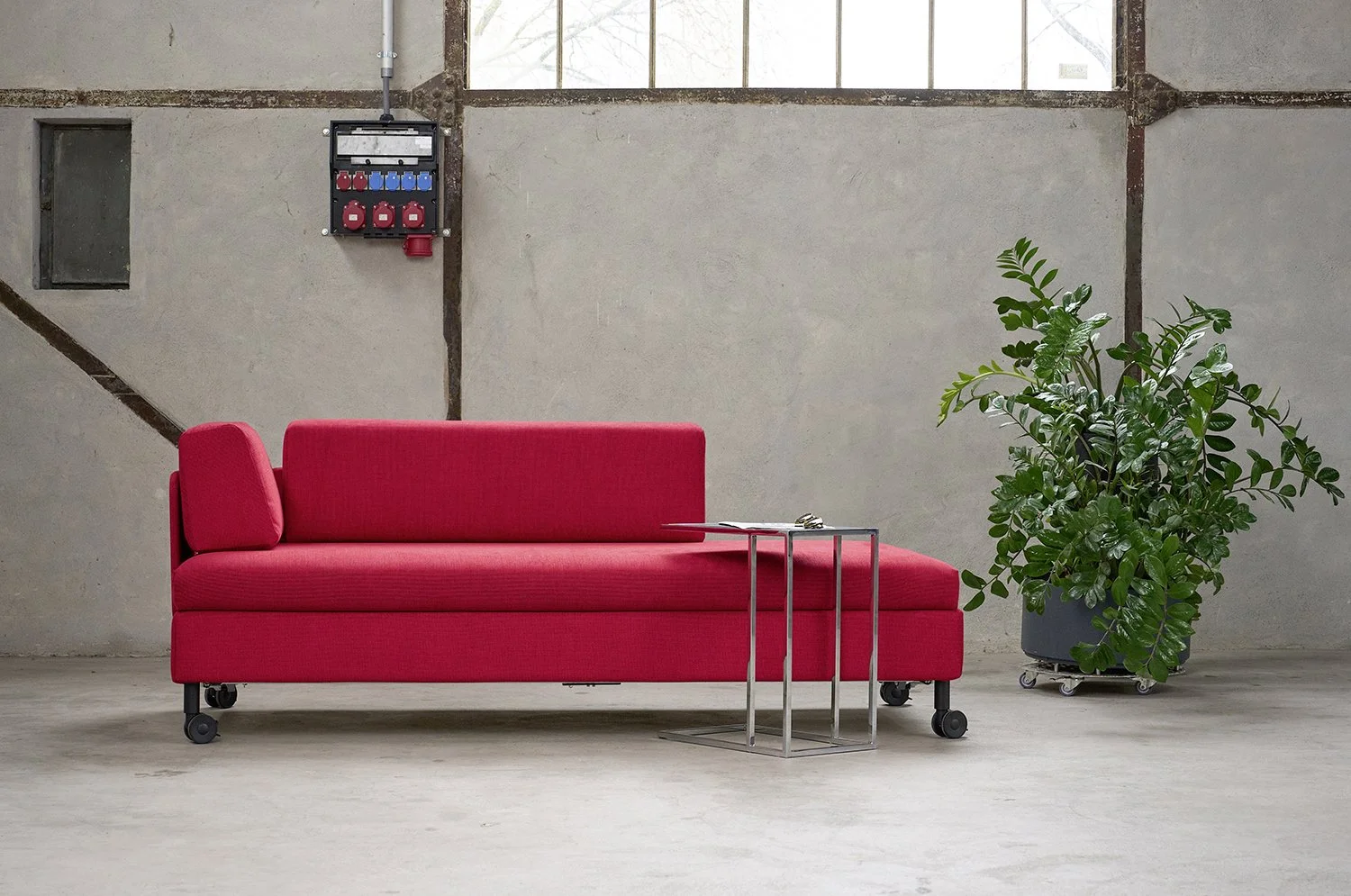 Ein rotes Sofa auf Rollen neben einem kleinen metallenen Beistelltisch vor einer Wand in einer Loft mit Fenstern und einer Pflanze in einem schwarzen Topf.