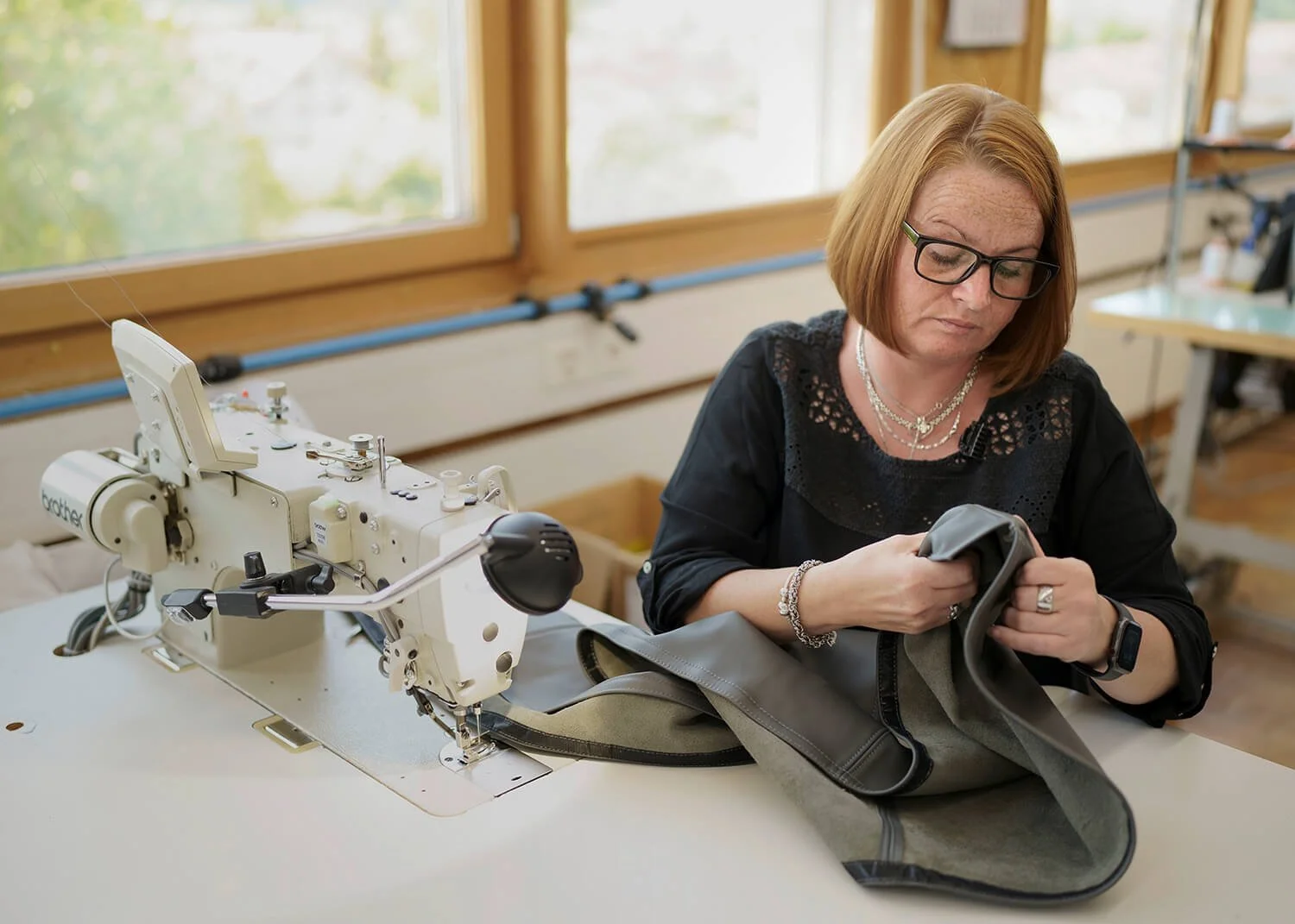 Corporate Portrait einer Frau an einer Nähmaschine. Sie näht einen Stoffbezug für ein Bettsofa mit Produktion in Zürich. Im Hintergrund sind große Fenster und Holzrahmen.