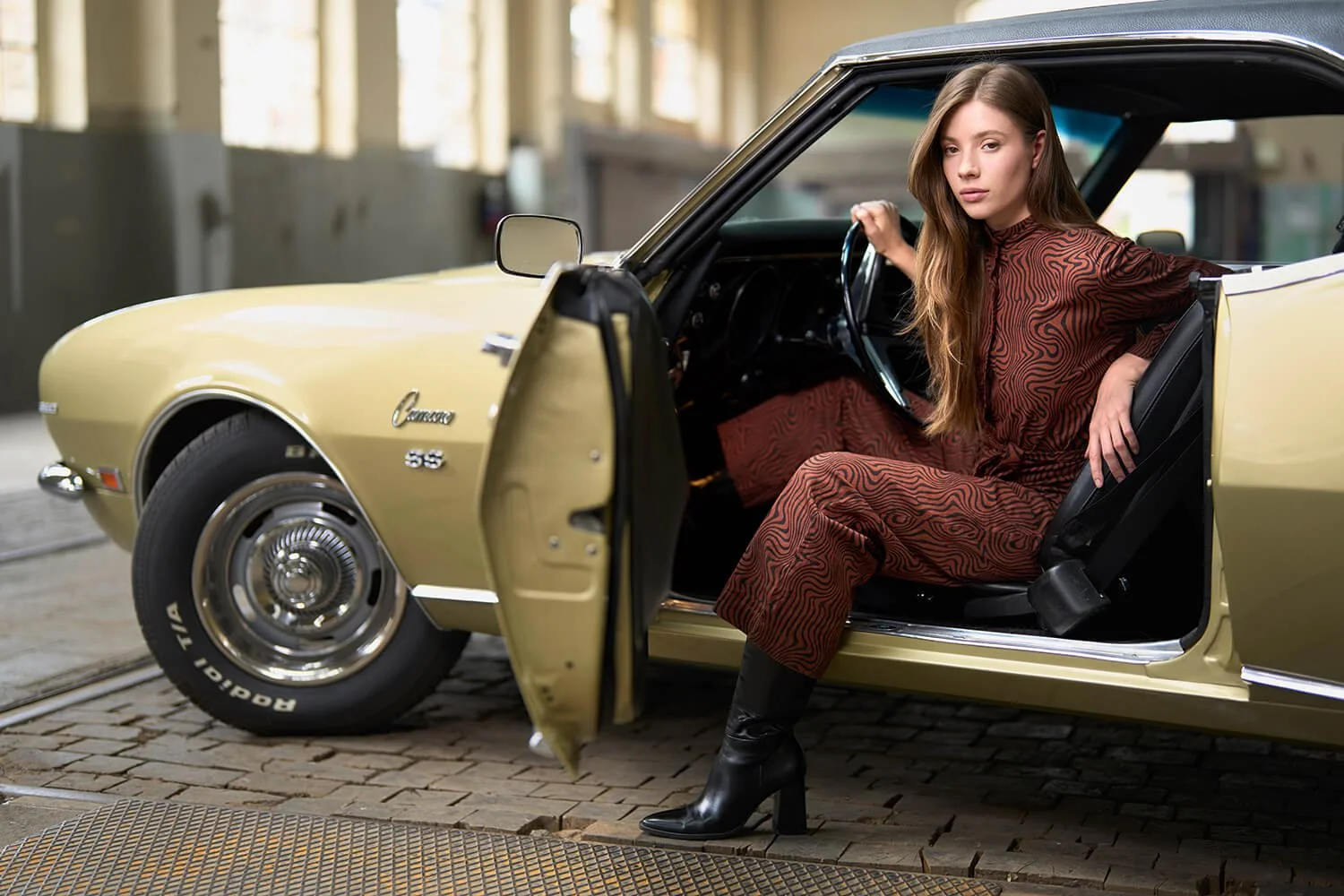 Modefotografie in einer grossen leeren Gewerbehalle. Eine junge Frau sitzt in einem goldenen Oldtimer-Cabriolet. Sie trägt einen braunen brauen Einteiler, mit gemustertem Outfit und schwarze Stiefel.