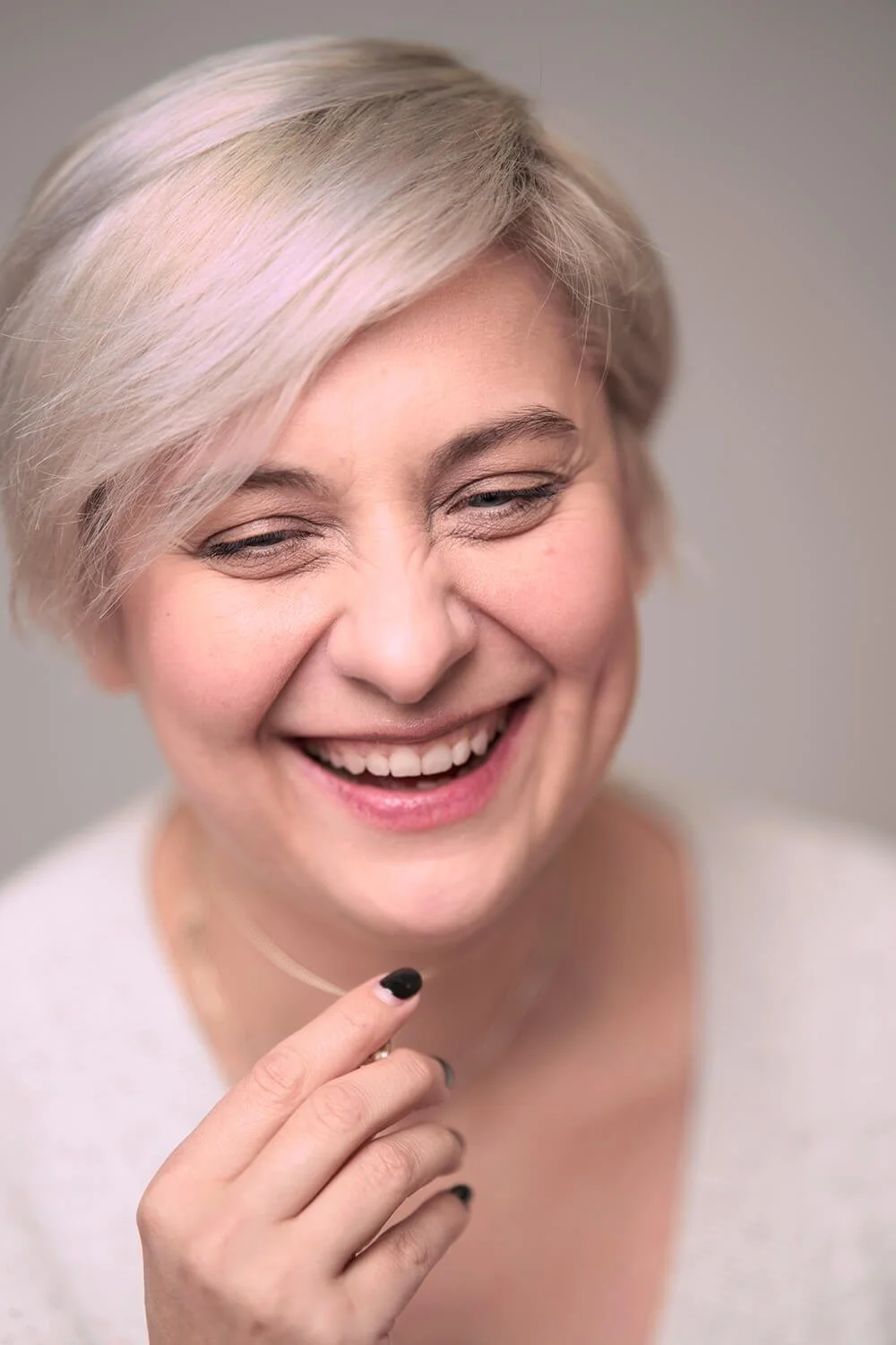 Fotgrafie einer lachenden Frau mit kurzen, platinblonden Haaren und dunklem Nagellack, lächelnd mit zusammengelegten Händen.