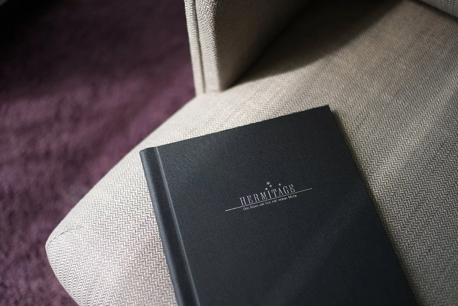 Fotografie eines eleganten Menüs mit silberner Schrift auf einem beige gepunkteten Sessel, im Hintergrund ein lila Teppich.