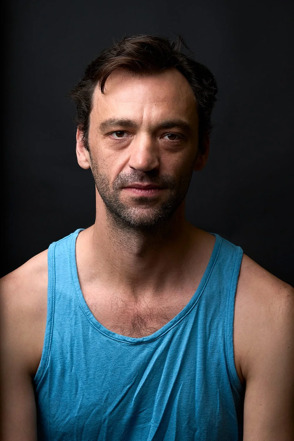 Porträt eines Mannes mit dunklem Haar, Dreitagebart und blauem Tanktop vor einem schwarzen Hintergrund.