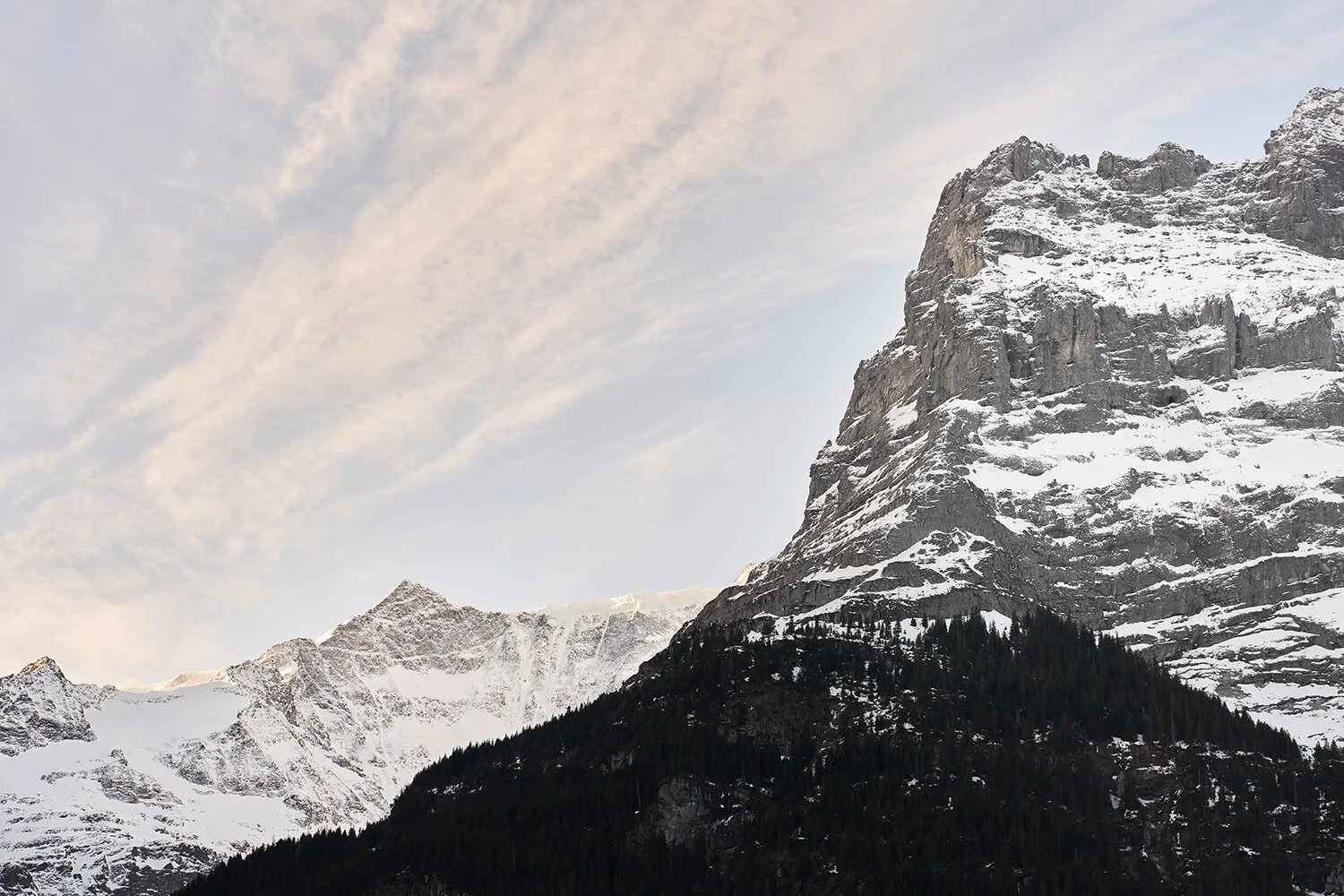 Stimmungsvolle Fotografie eines schneebedeckten Bergs mit dunklem Wald im Vordergrund, blauer Himmel mit Wolken