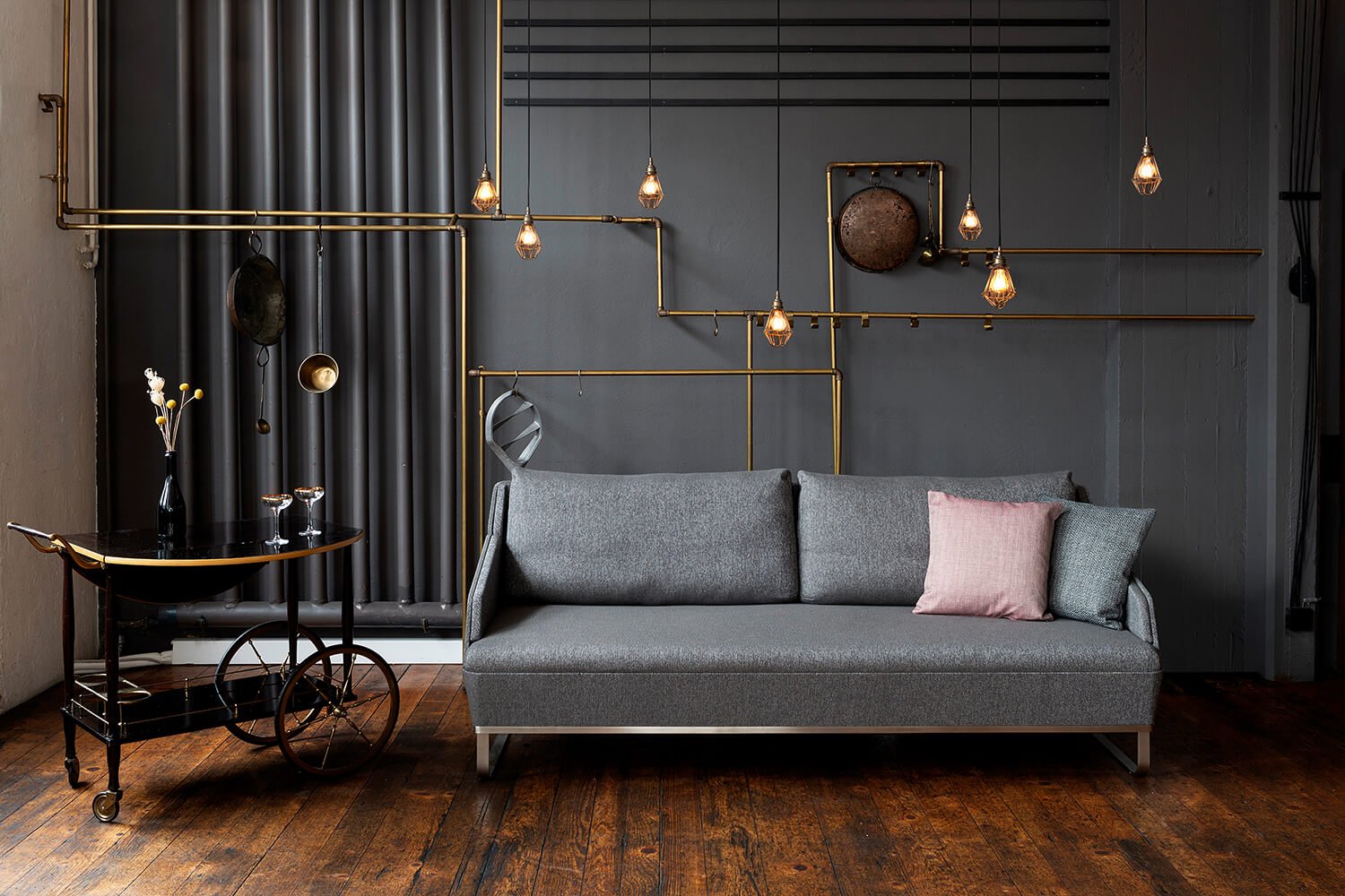 Interior Fotografie eines modernesn Wohnzimmers mit grauem Sofa, hellem Kissen, dunkler Wand, goldenen Kabelleuchten, dunklem Vorhang, dunklem Holzboden, vintage Barwagen, kleinerm Tisch mit Wein, dekorativen Gegenständen, Lampen, Accessoires.
