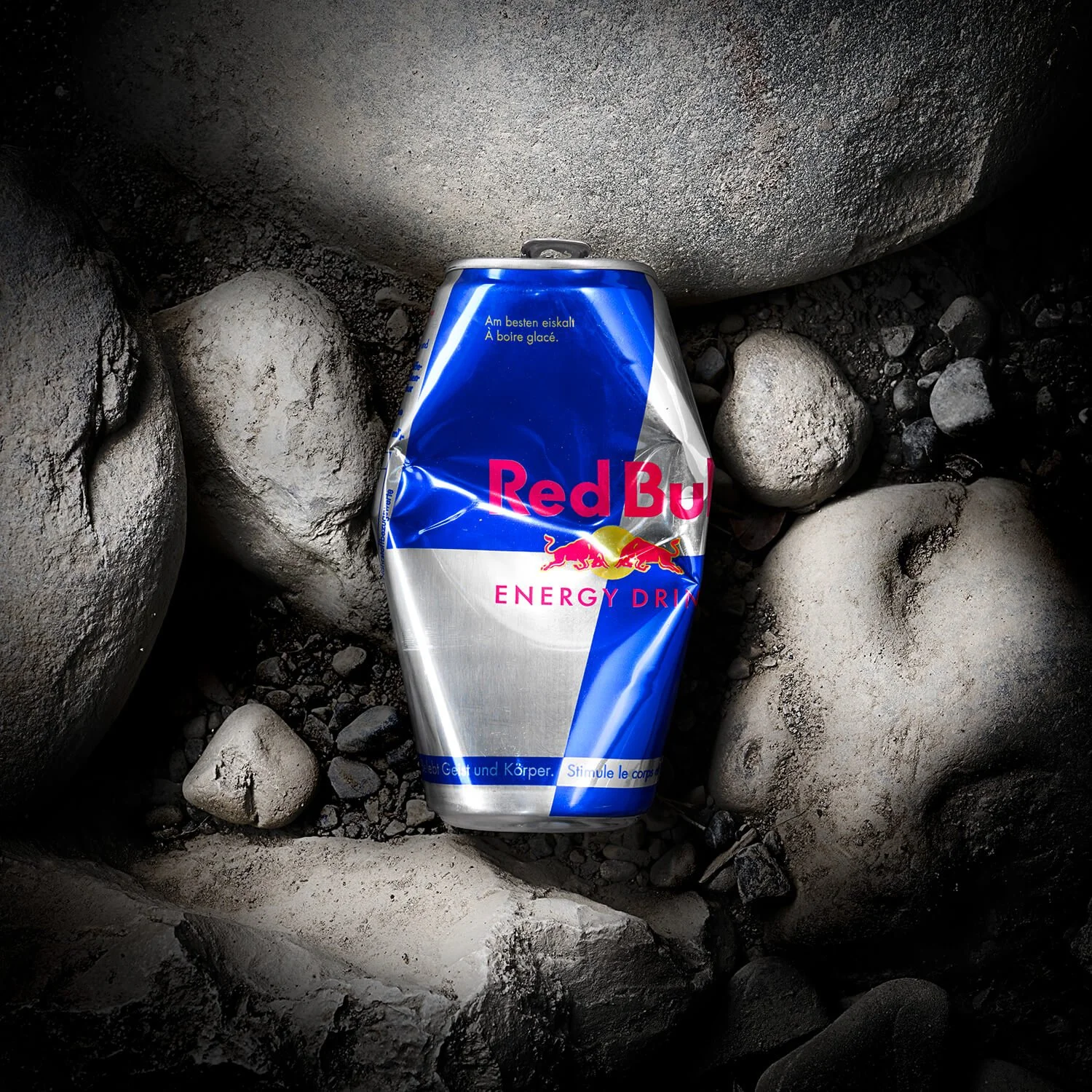 Eine wie eine Werbefotografie wirkende Darstellung einer zerquetschten Dose Red Bull Energy Drink, die weggeworfen auf einem Boden aus Steinen und Sand liegt.