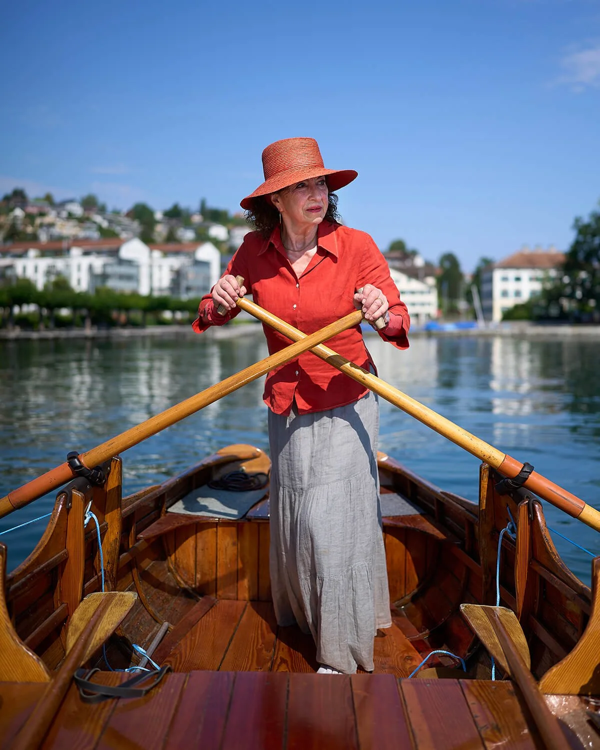 Stilvolle Fotografie einer älteren Frau auf einem Holzboot, die mit Ruder paddelt, einen Strohhut trägt und eine rote Bluse. Im Hintergrund sind Häuser und Bäume am Ufer eines Flusses bei schönem Wetter zu sehen.