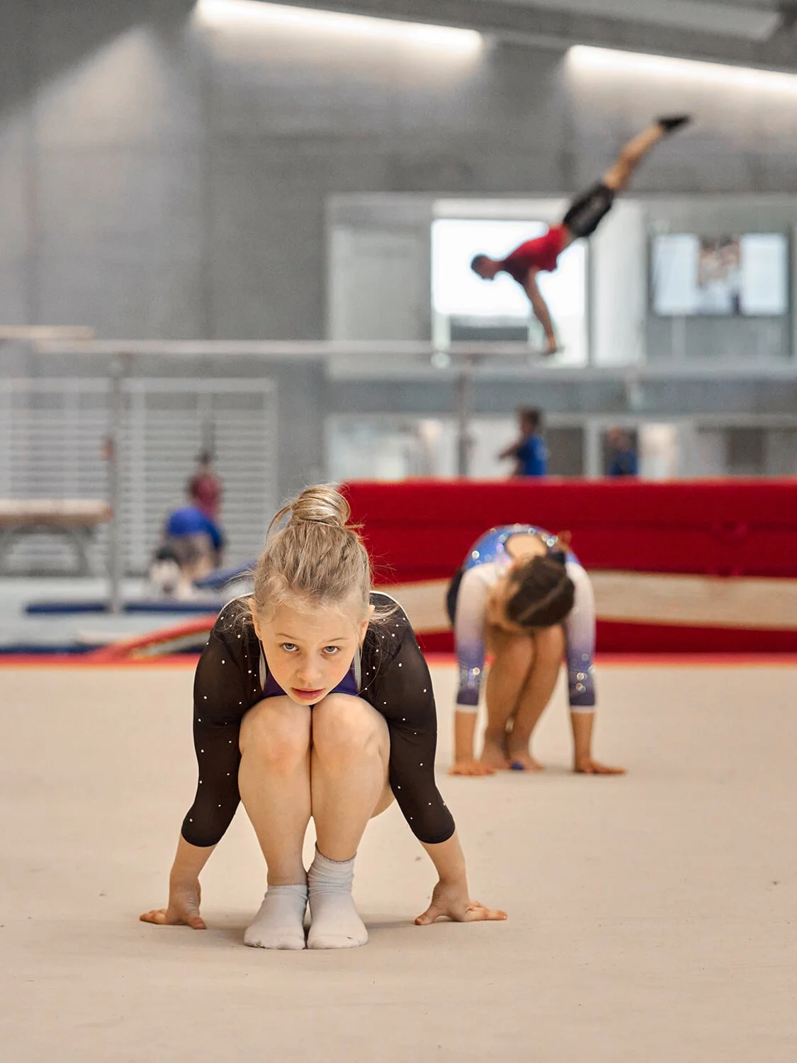 Fotografische Komposition einer jungen Turnerin in einer Sporthalle, die auf dem Boden kauert und einem Turner im Hintergrund, der eine Acrobatic-Übung am Barren ausführt.