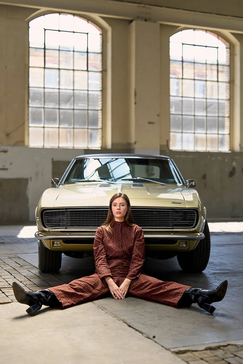 Modefotografie in einer grossen leeren Gewerbehalle. Eine junge Frau in einem braunen Einteiler und schwarzen Stiefeln sitzt auf dem Boden vor einem klassischen Oldtimer in einem industrialähnlichen Raum mit großen Fenstern.