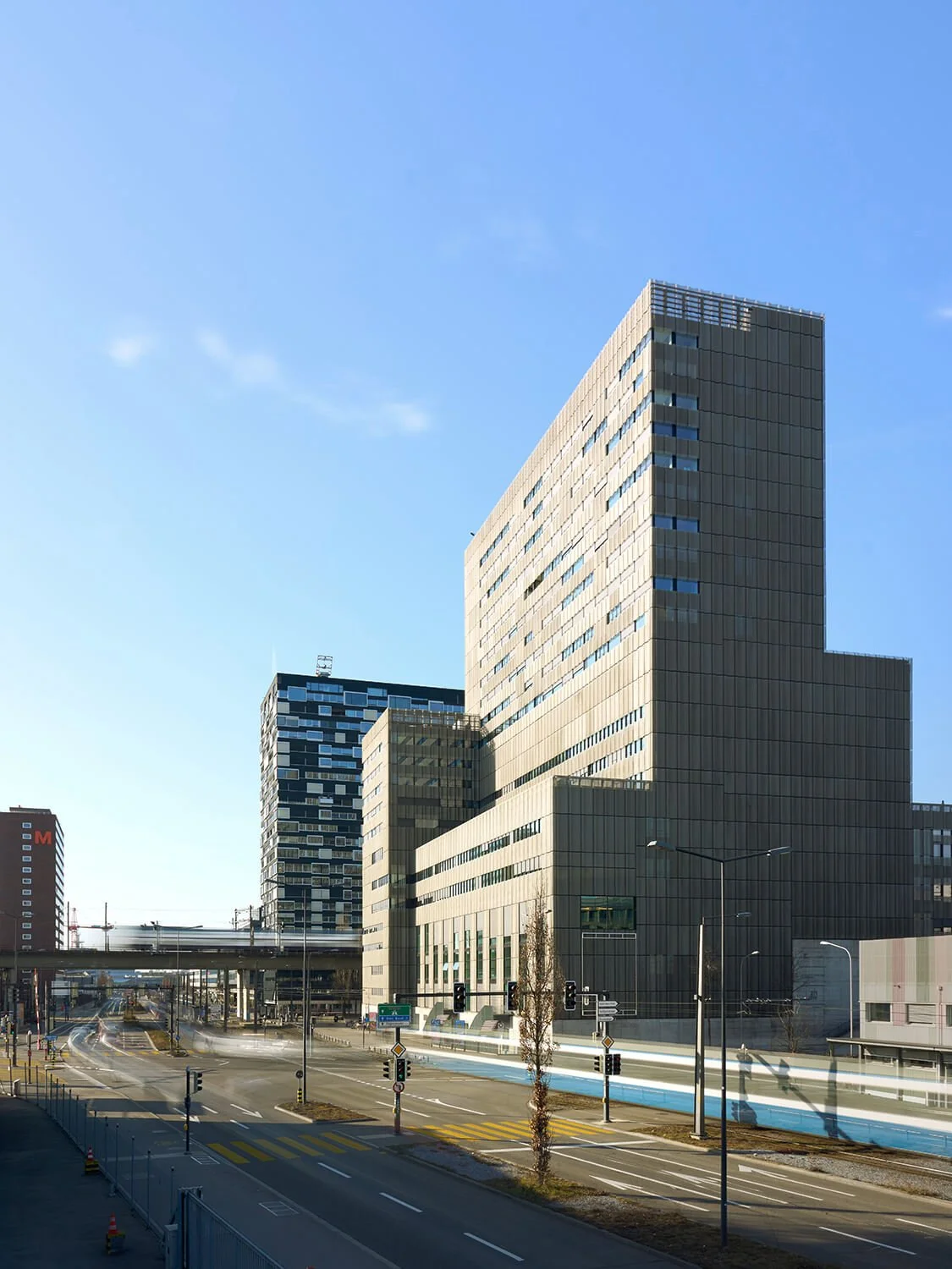 Architekturfotografie mit Langzeitaufnahme der ZHdK im Toni Areal in Zürich bei Tageslicht, mit klarem blauen Himmel, Straßen und Verkehrssignale im Vordergrund.