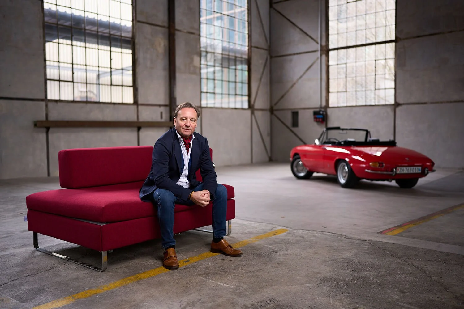 Ein Mann sitzt auf einer roten Couch in einer Industriehalle neben einem roten Oldtimer-Cabriolet. Im Hintergrund sind große Fenster und die Wände bestehen aus rohen Betonplatten.