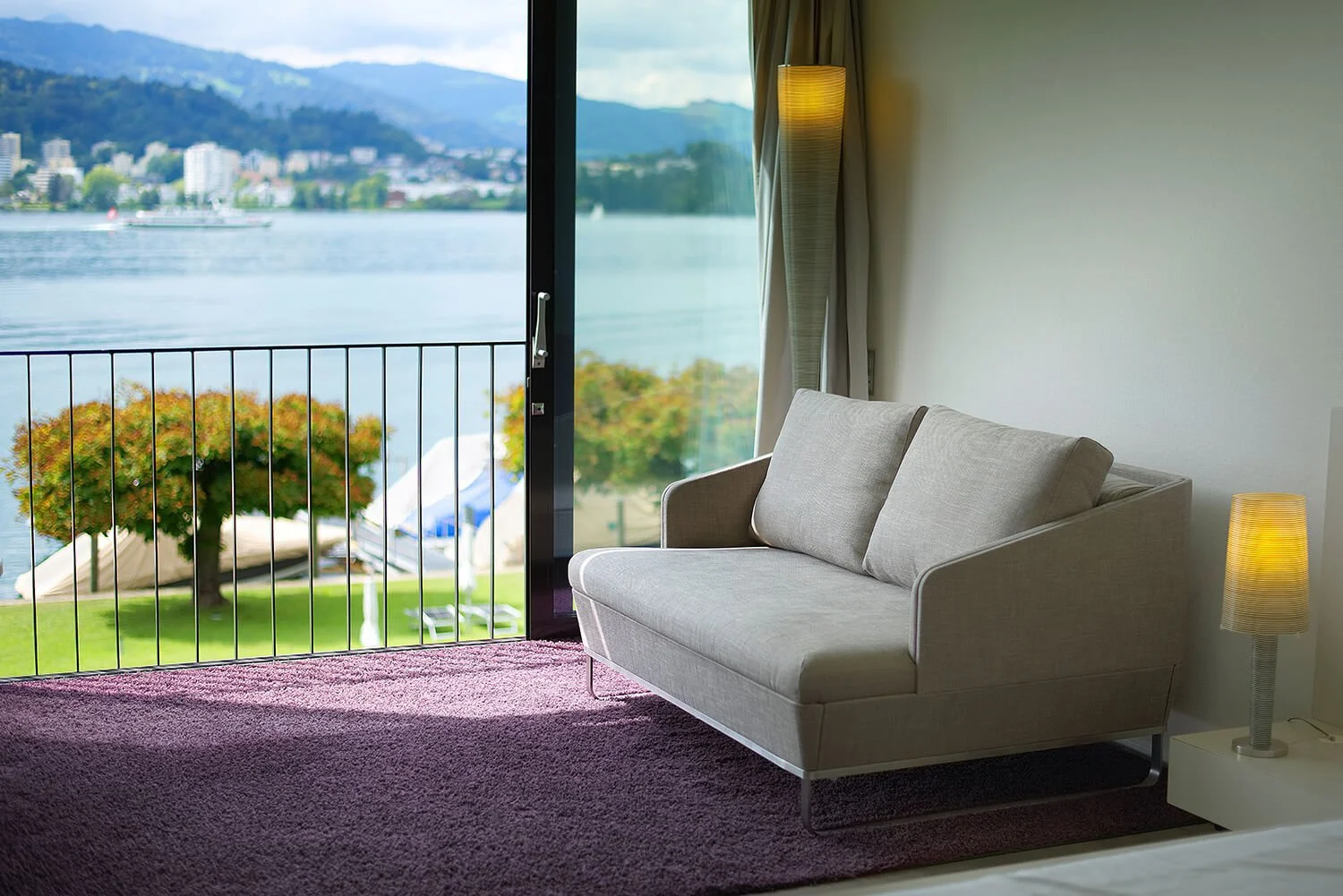 Interiorfotografie eines Innenraums eines Zimmers mit grauem Sofa, neben einem Nachttisch mit Lampe, offenem Balkon mit Blick auf einen Fluss und eine Stadt im Hintergrund