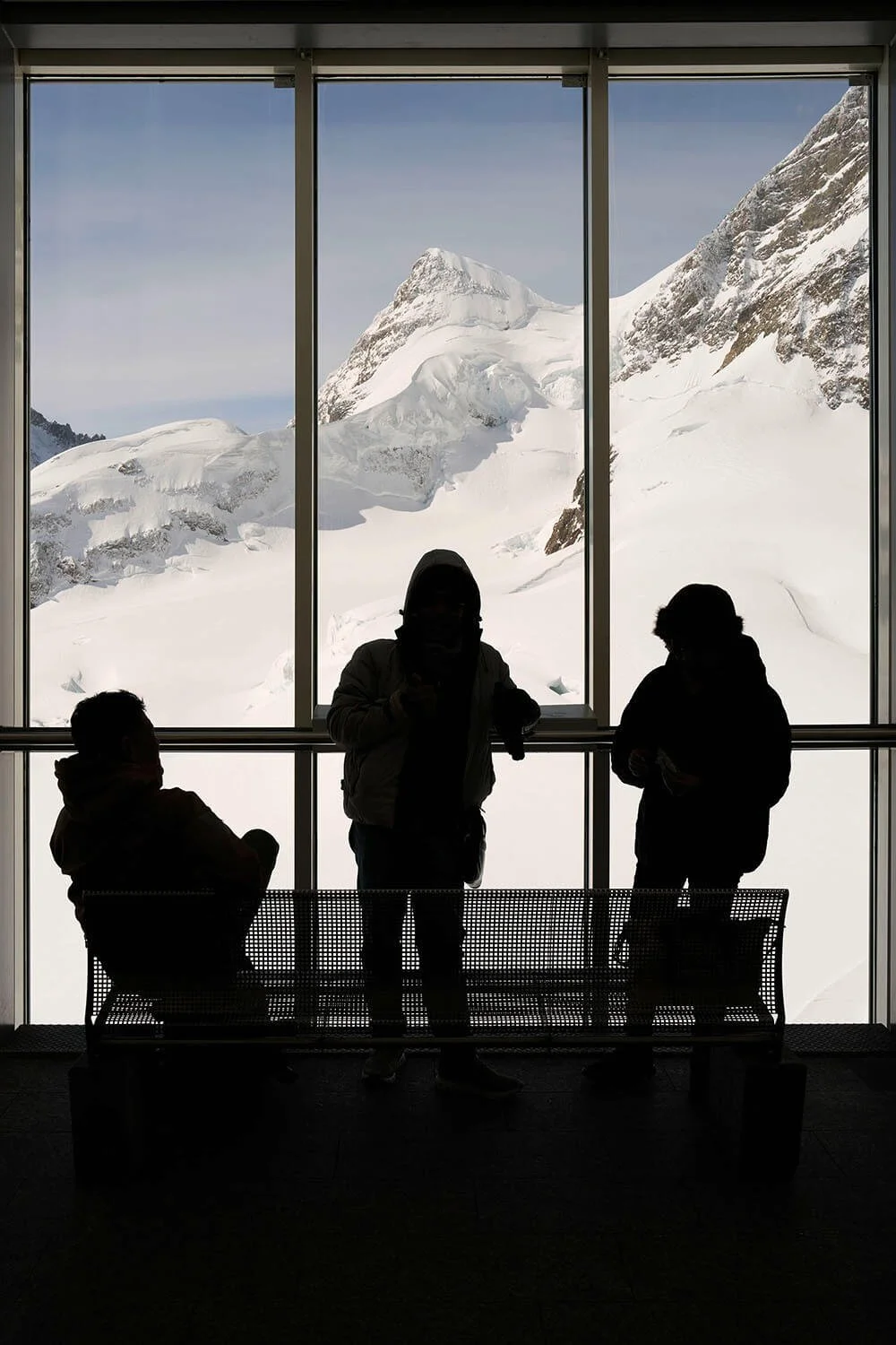 Abstrakte Fotografie von drei silhouettierten Personen, die vor einem grossent Fenster sitzen und stehen, das einen verschneiten Berg mit Gletschern und Felsen zeigt.