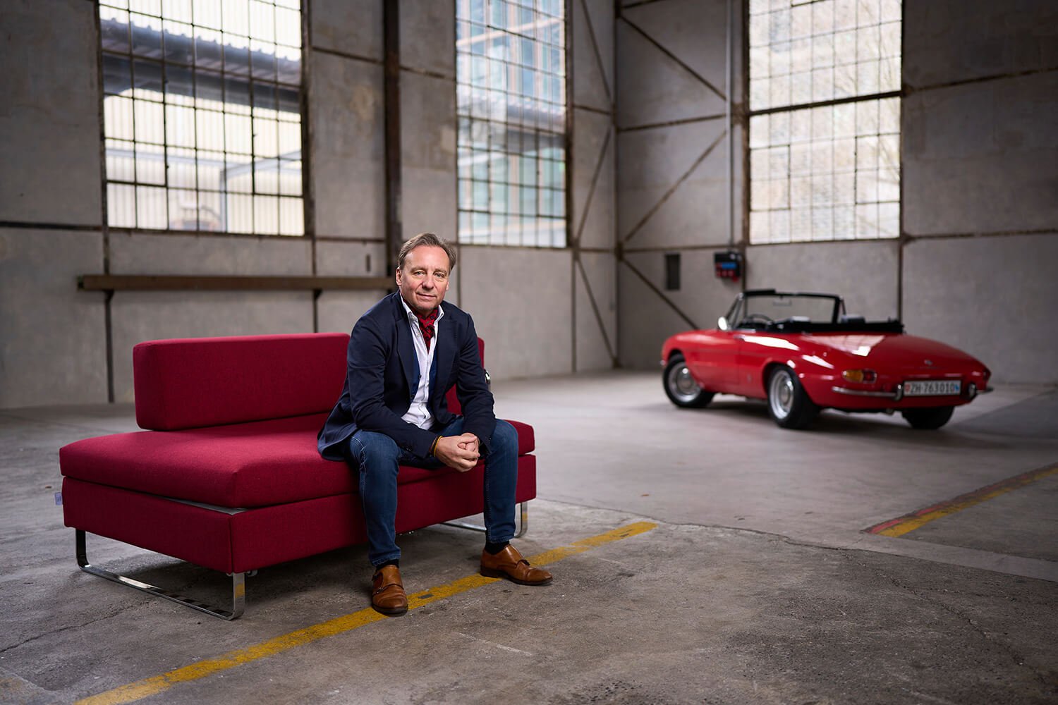 Corporate Portrait eines Mannes, der auf einem roten Bettsofa der Marke BED for LIVING sitzt. Er befindet sich in einer großen, industriellen Halle mit hohen Fenstern. Im Hintergrund steht ein rotes Cabrio-Auto.