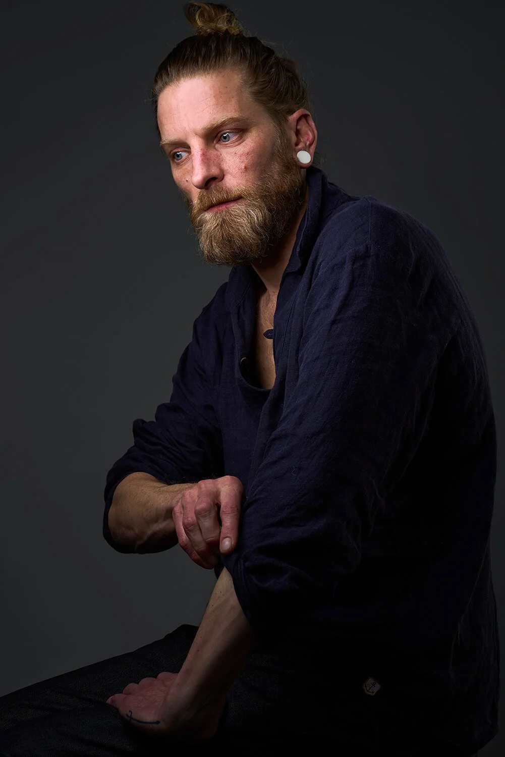 Portraitfotografie eines Mannes mit braunem Bart, langer Haarsträhne in einem Dutt. Er trägt ein schwarzes Shirt und eine dunkle Jacke, sitzt vor dunklem Hintergrund, schaut nach unten, berührt seinen rechten Unterarm.