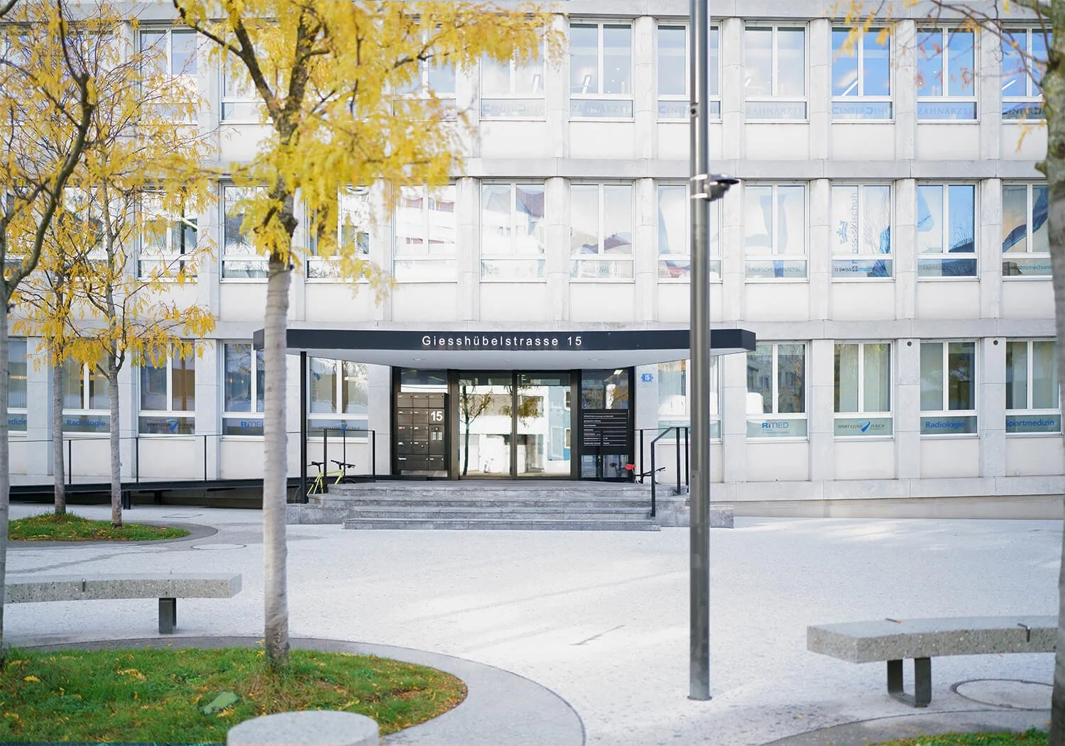 Bürogebäude mit dem Eingangsbereich an der Giesshübelstrasse 15 in Zürich, umgeben von Bäumen mit gelbem Herbstlaub, einige Fahrräder vor der Tür, es ist Tag mit bewölktem Himmel. Hier ist der Eingang zum Fotostudio von Samuel Köppel.