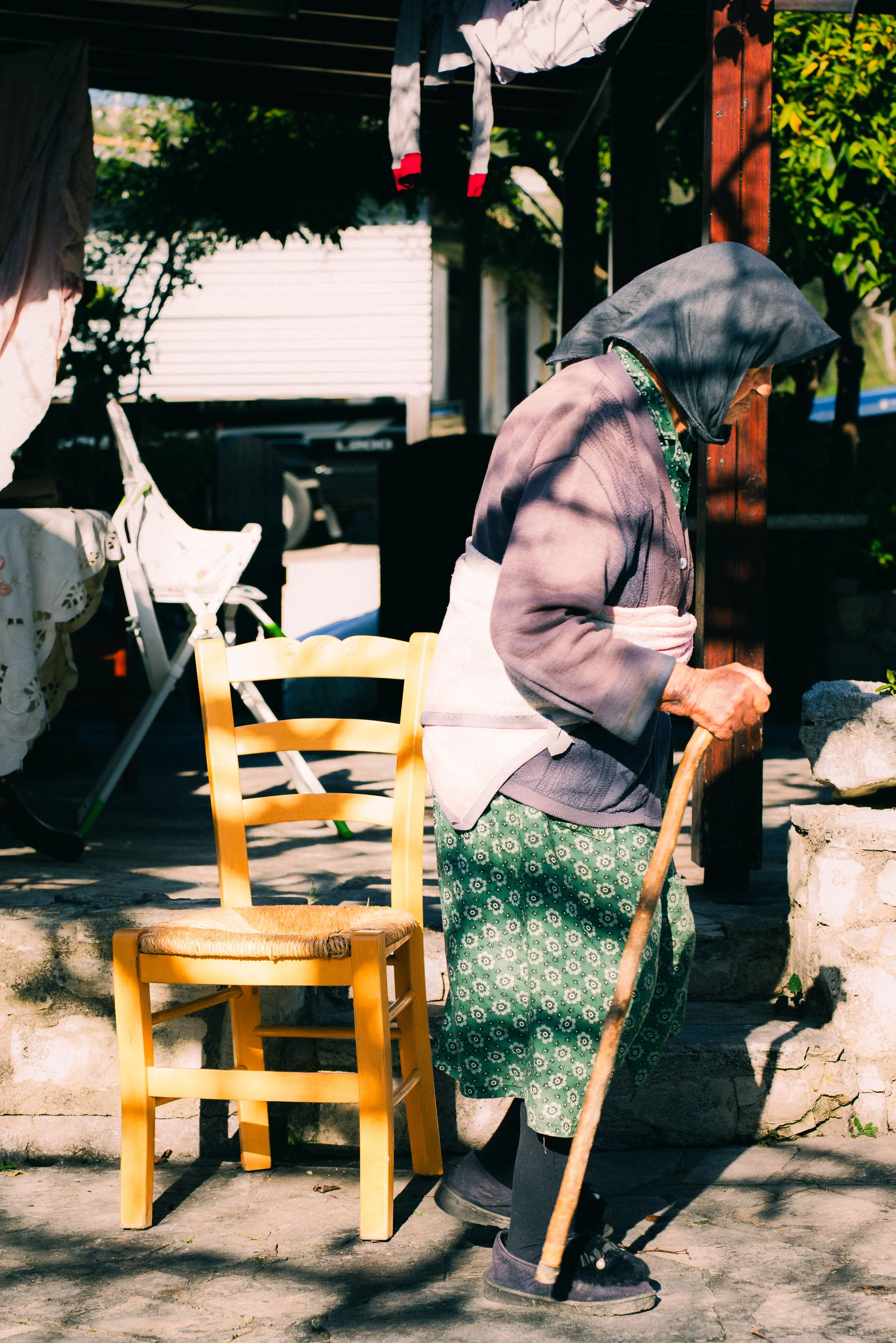 An elderly woman with a gray headscarf and patterned green pants stands outside, leaning on a cane next to a yellow wooden chair, in a shaded area with various household items and greenery around.