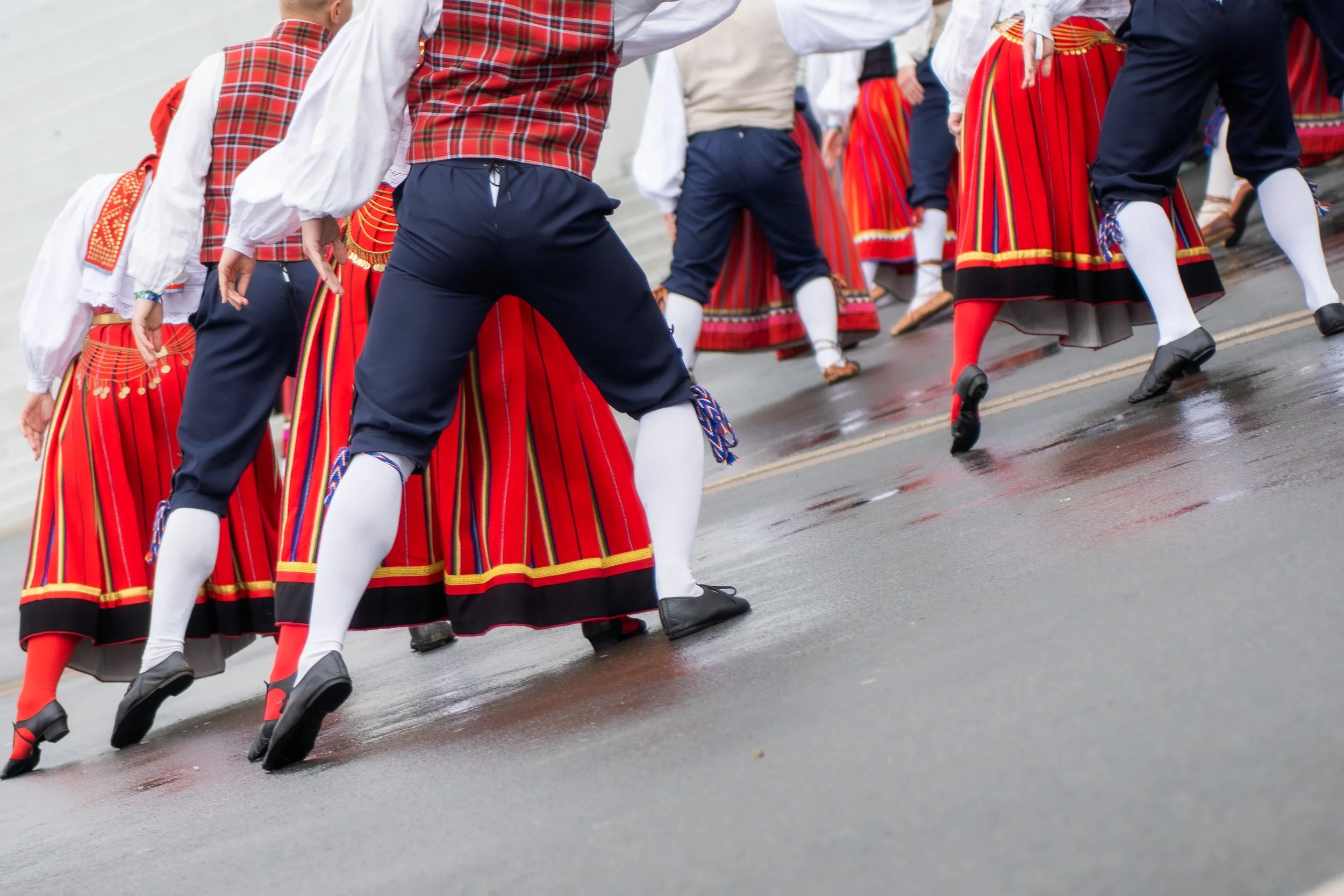 People in traditional colorful folk costumes dancing during a cultural parade on a wet street.