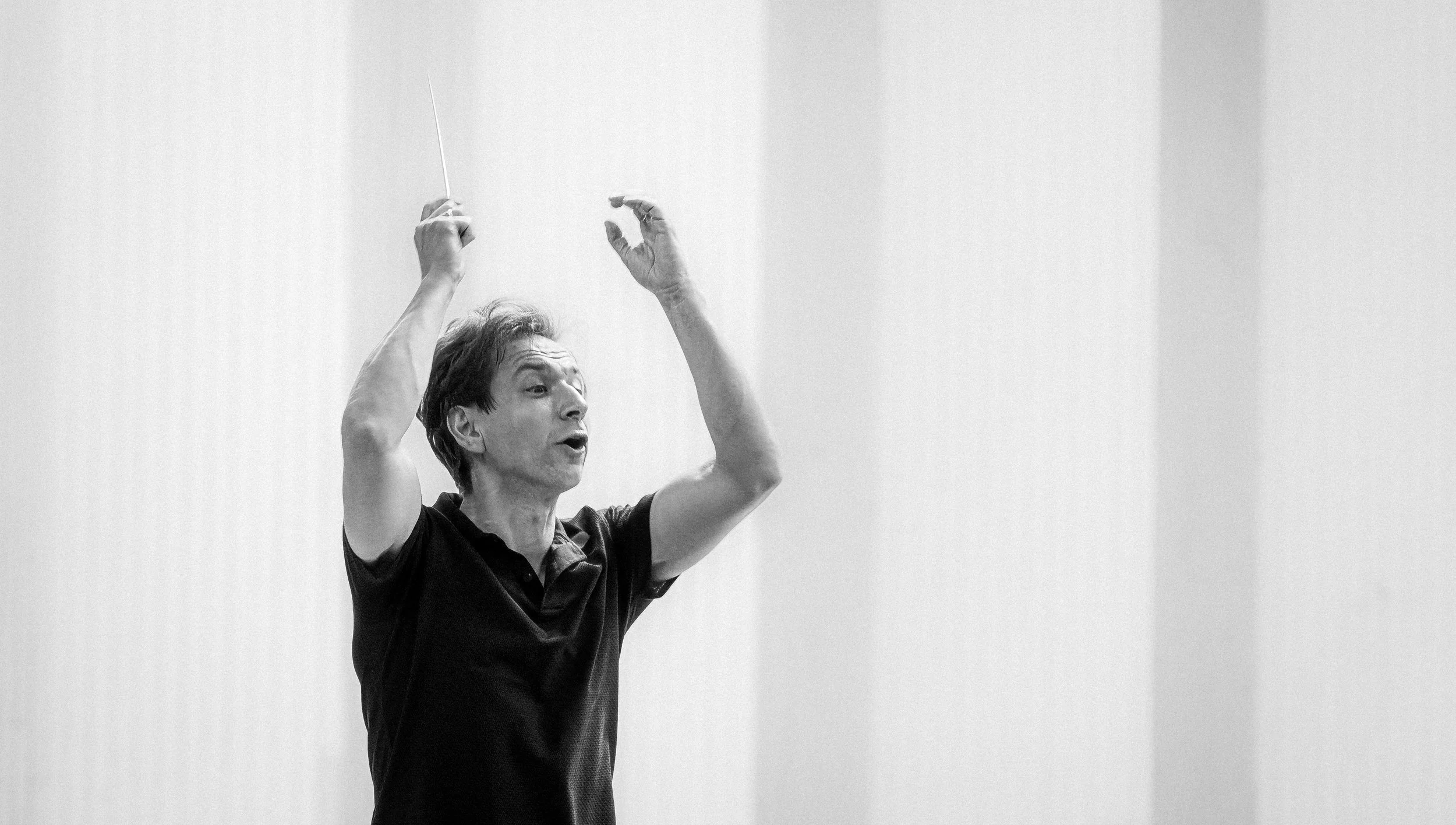 A man with dark hair conducting an orchestra with a baton, expressing concentration, in a black and white photo.