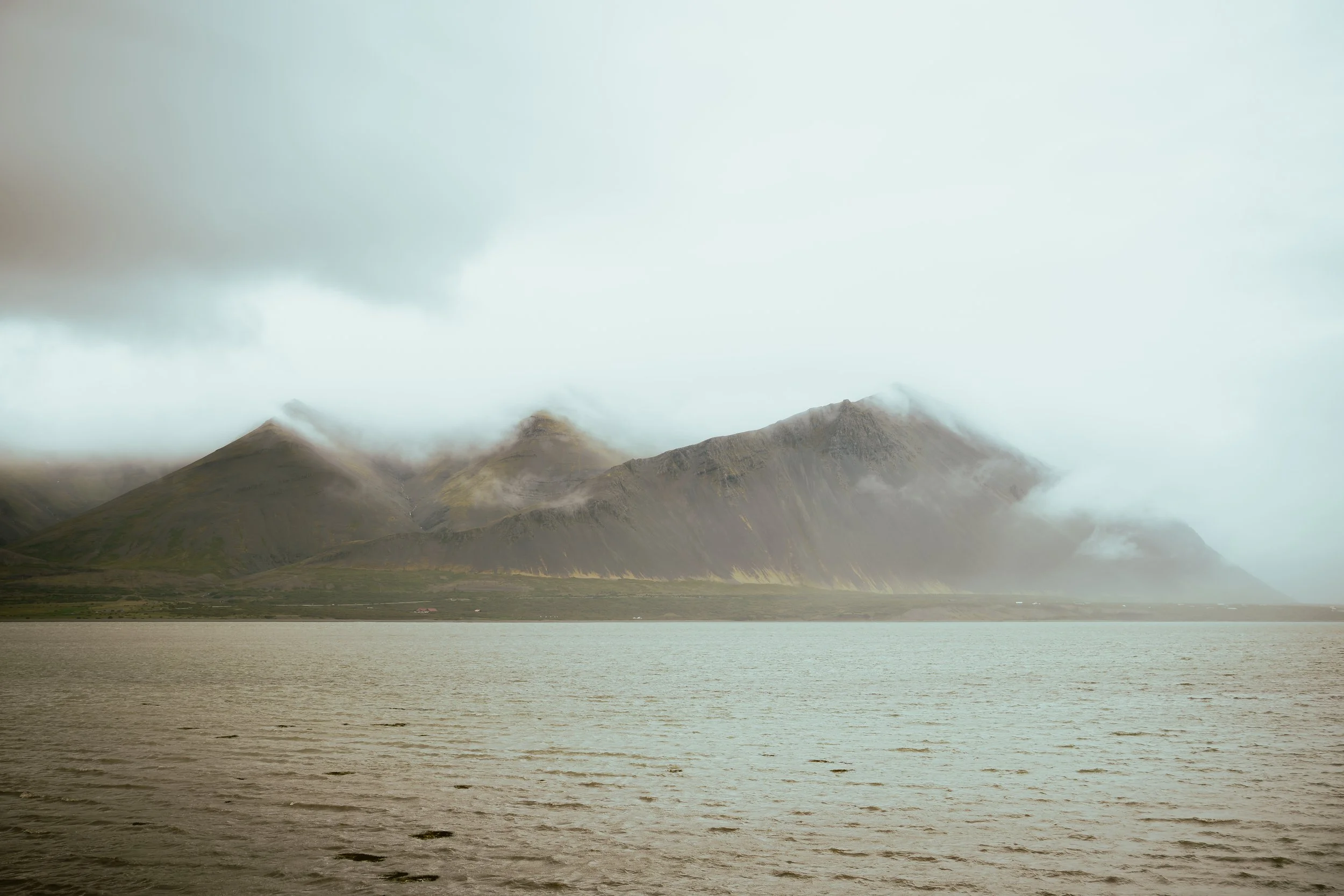 Mountains with clouds and fog near a body of water, possibly a lake or ocean, under an overcast sky.