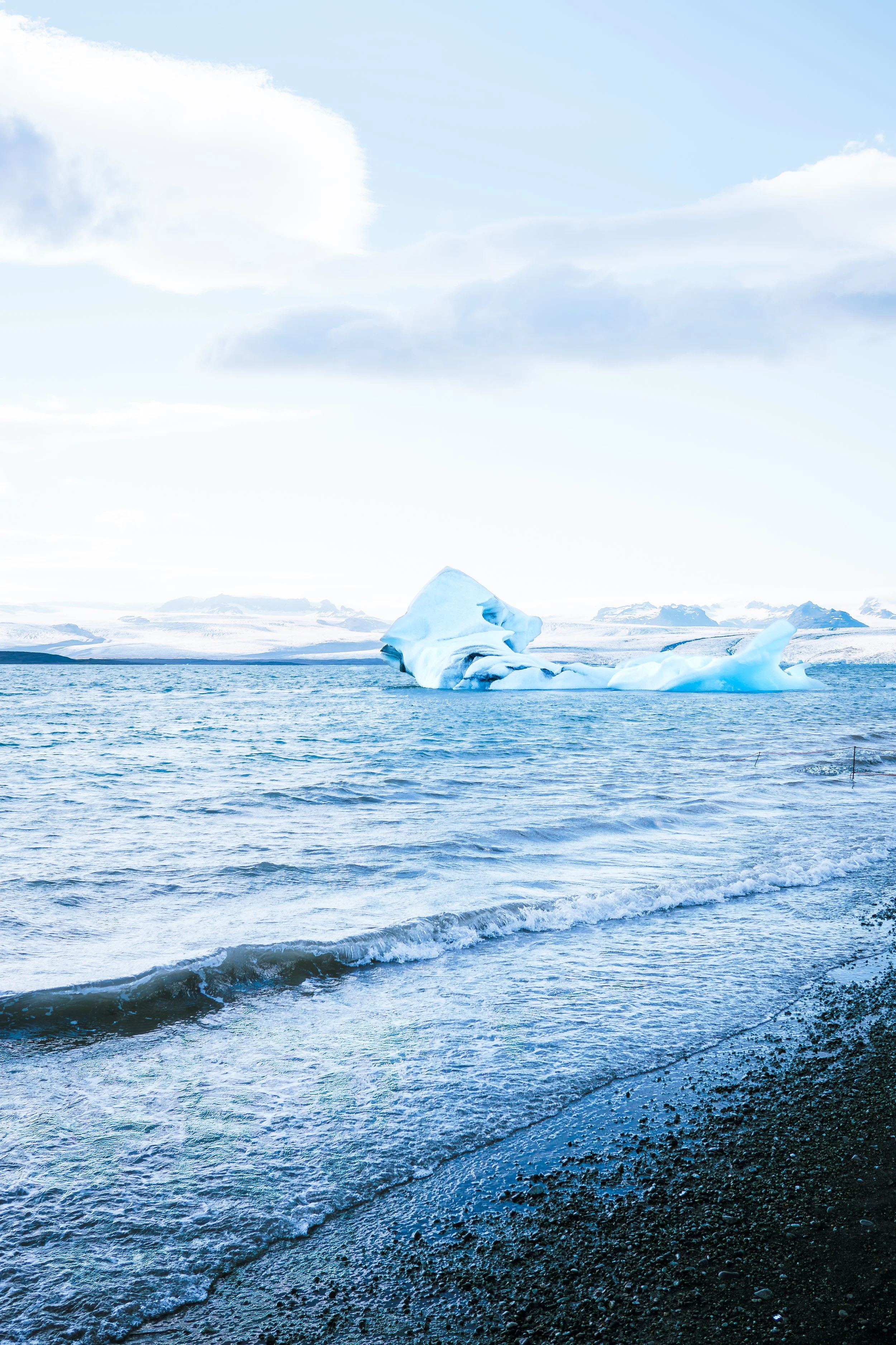 Cold ocean water with a large iceberg floating and snow-covered mountains in the background under a cloudy sky.