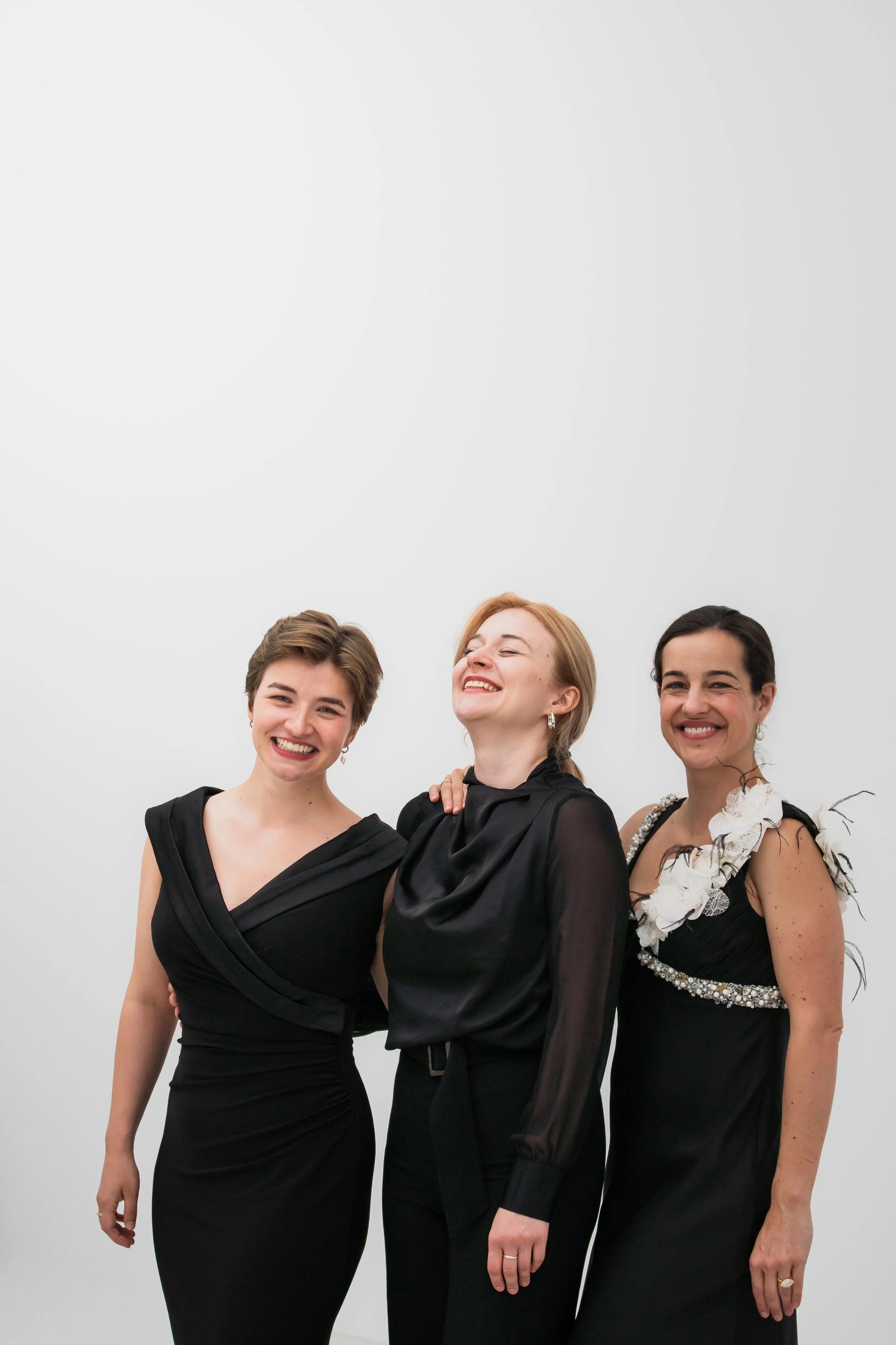 Three women dressed in elegant black dresses smiling and standing together against a plain gray background.