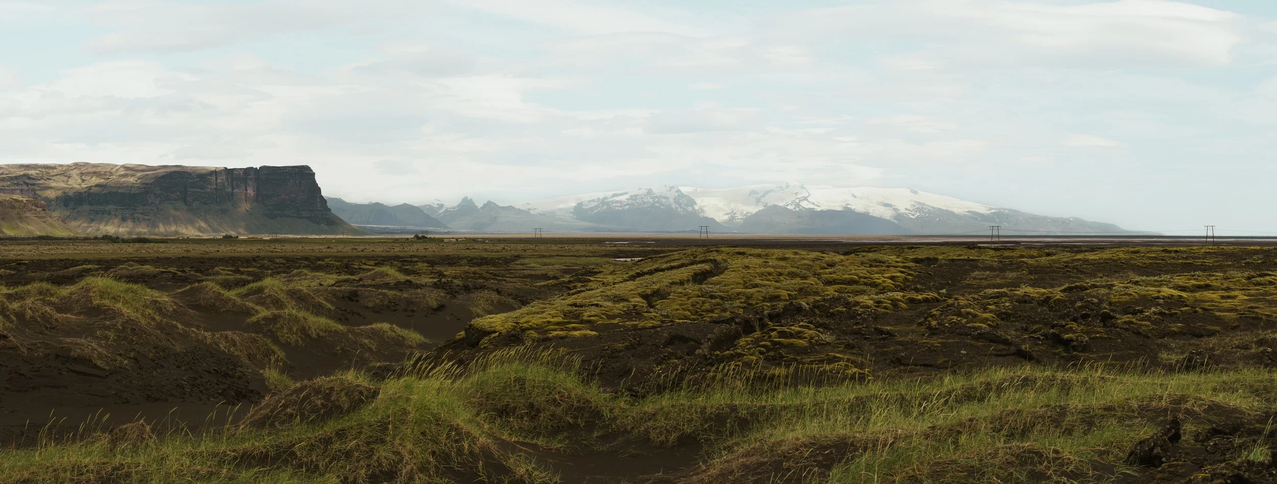 A wide landscape view of a volcanic terrain with dark volcanic soil and patches of green grass, with mountains and snow-capped peaks in the background under a partly cloudy sky.