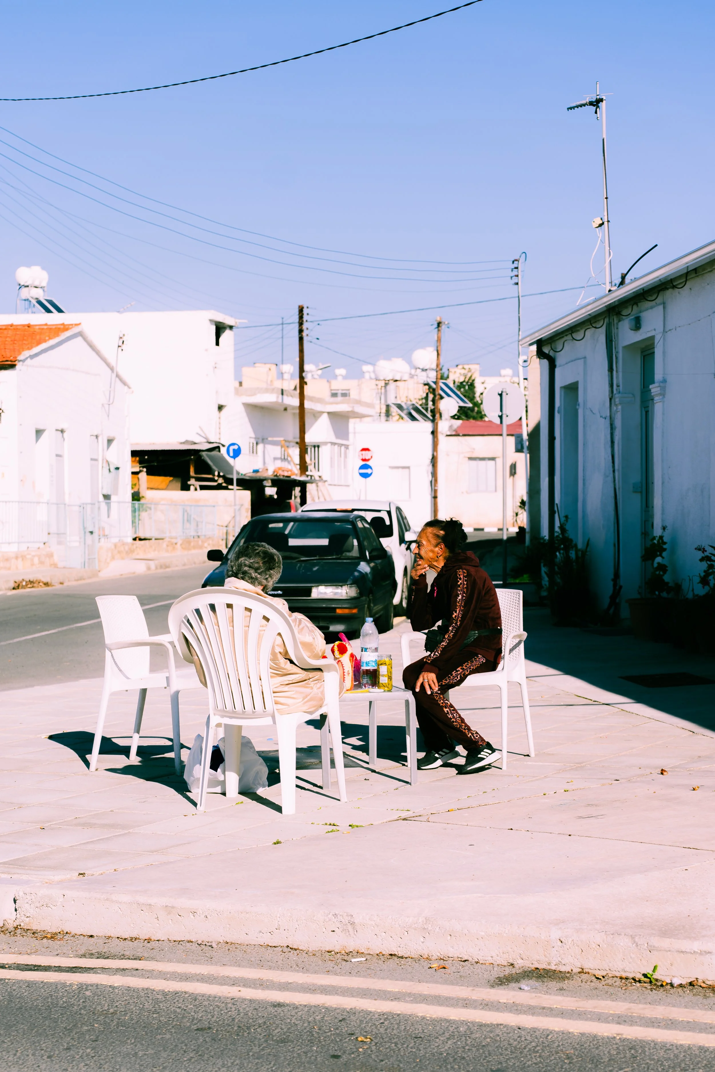 Two elderly women sitting at a white outdoor table on a sidewalk, engaged in conversation, with one woman wearing a beige coat and the other in a dark tracksuit. There are plastic chairs, a water bottle, and other small items on the table. The backgr