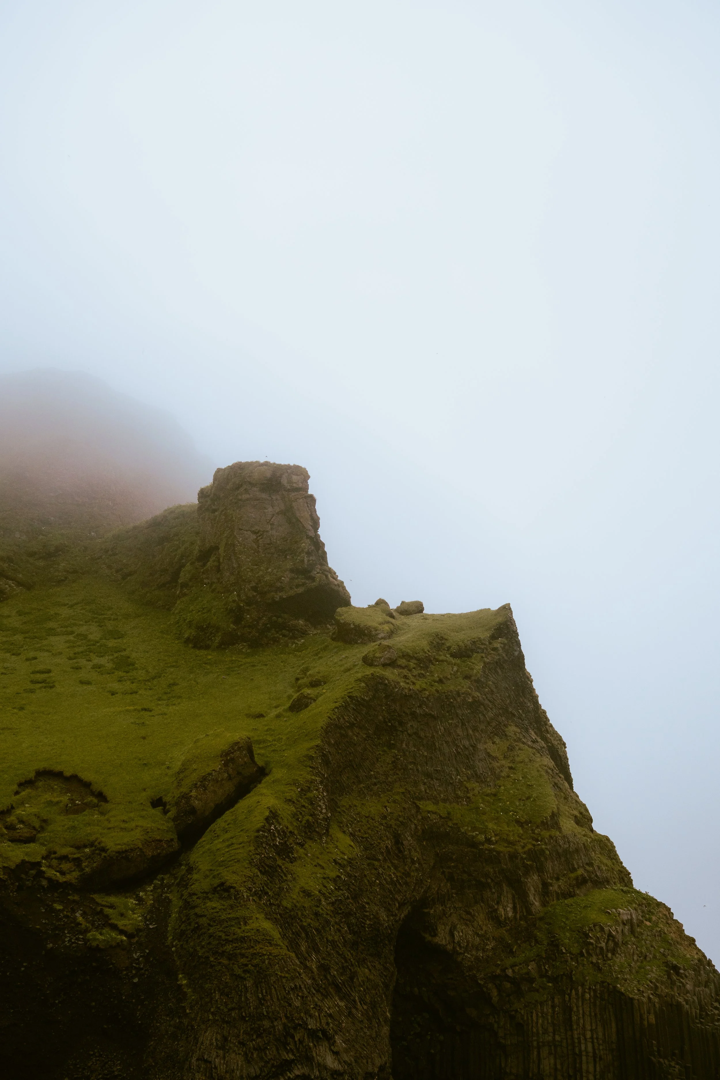Cliffside landscape with steep moss-covered rocks and foggy sky.