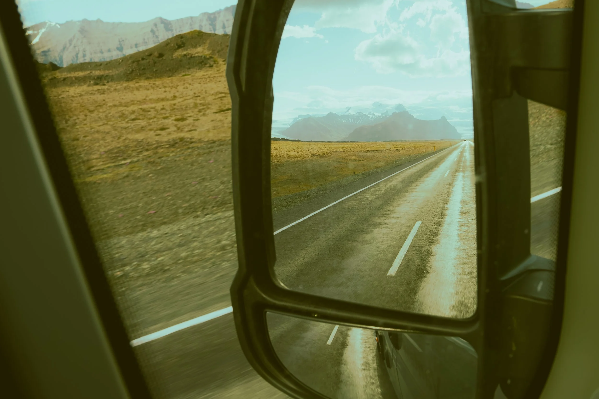 View from inside a helicopter showing a straight two-lane road passing through a barren landscape with mountains in the distance, under a partly cloudy sky.