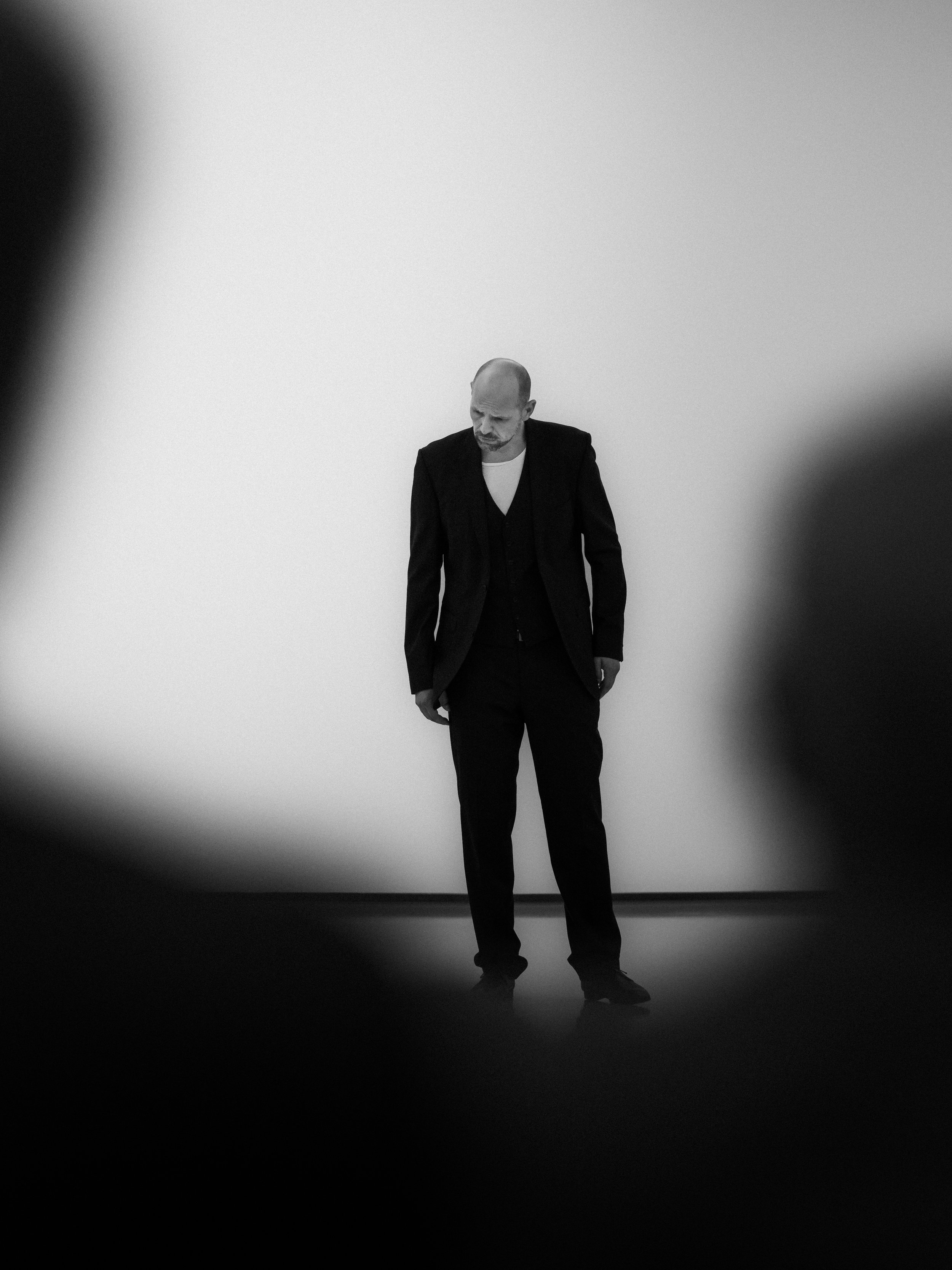 A black and white photo of a man with a shaved head wearing a dark suit and white shirt, walking on a plain background with his head down.