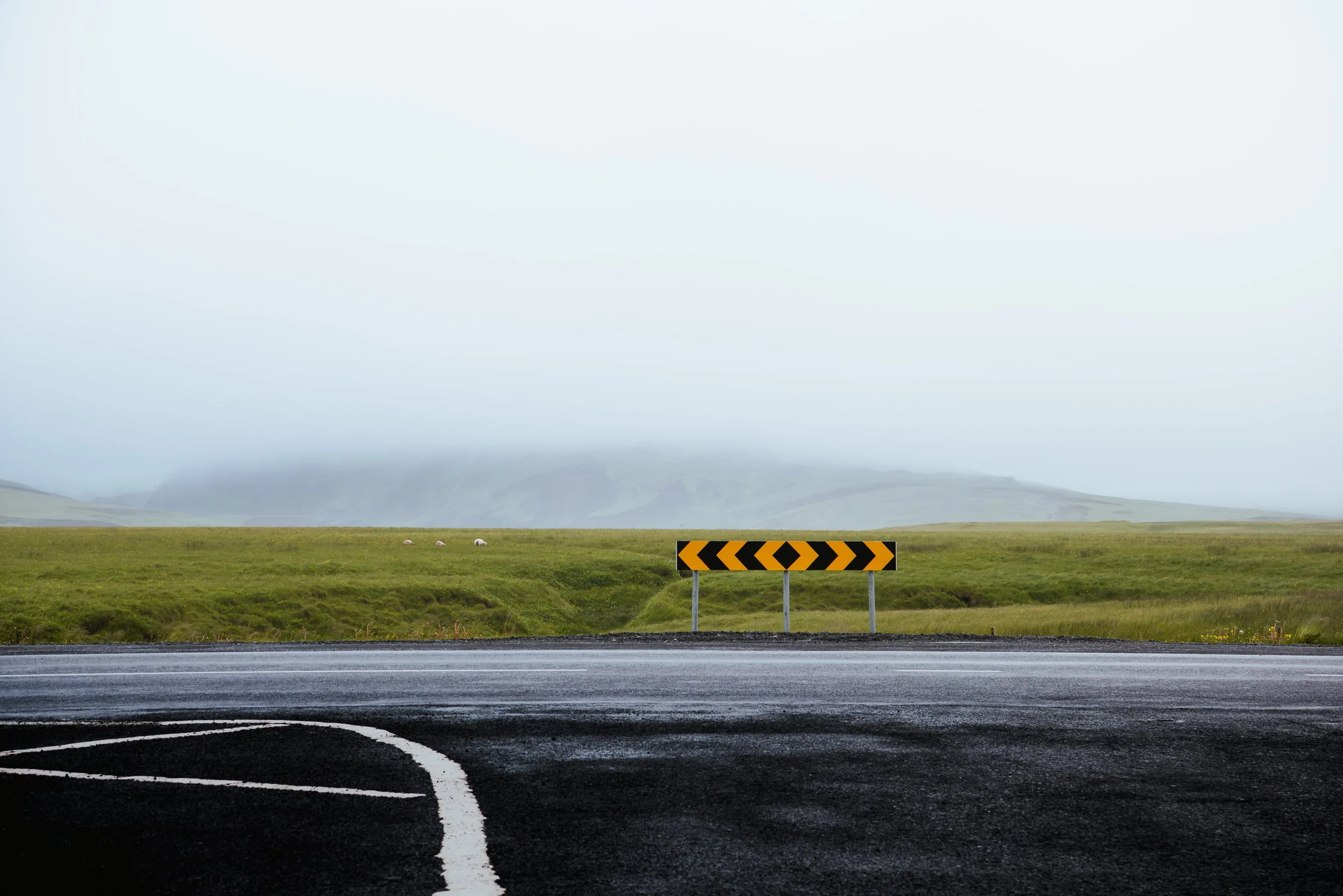 A two-lane road with a dark surface and white lane markings, including a curved arrow, in front of a grassy field and distant rolling hills, with a foggy sky overhead.