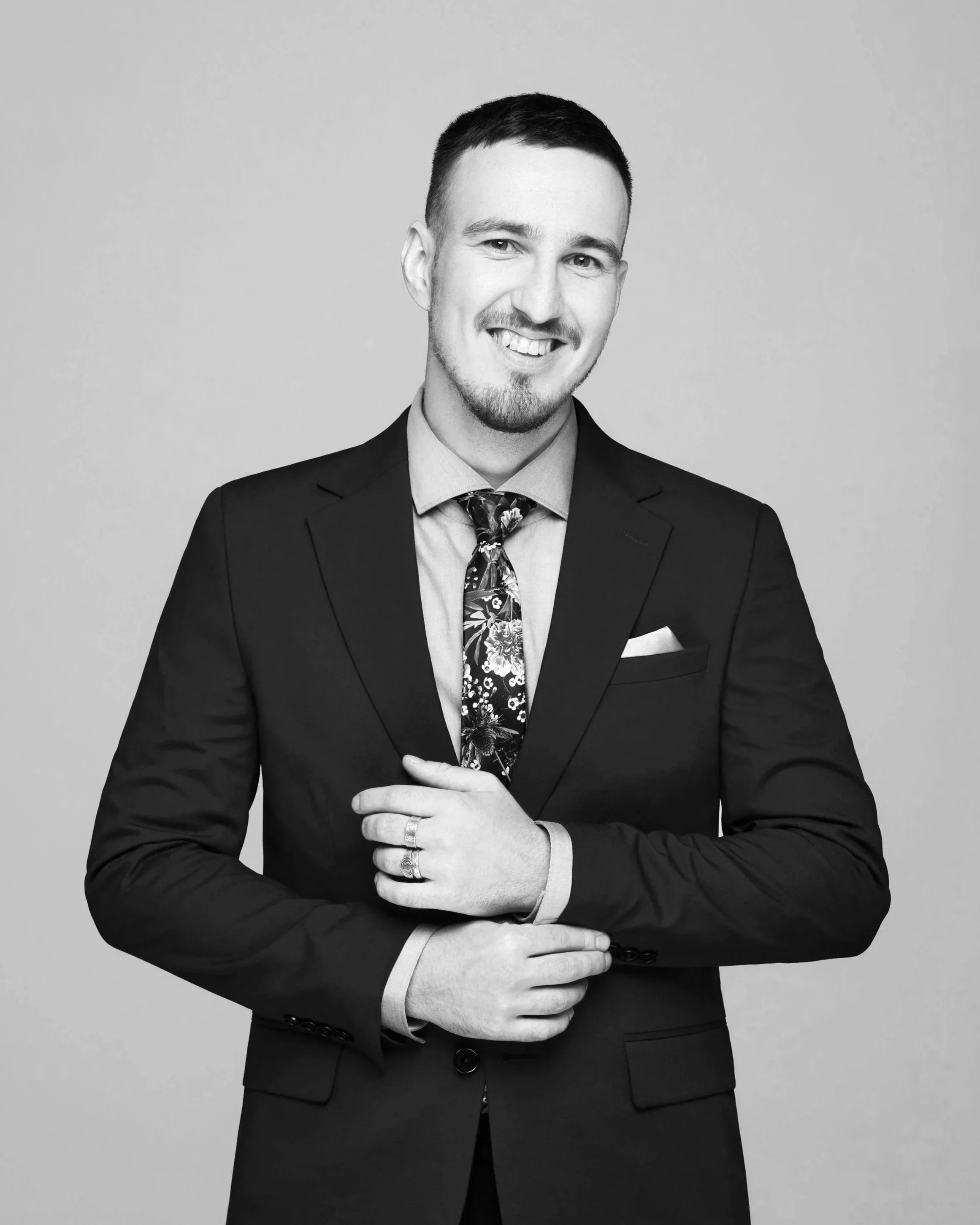 Black and white portrait of a smiling man in a suit with his arms crossed, wearing rings and a floral tie.