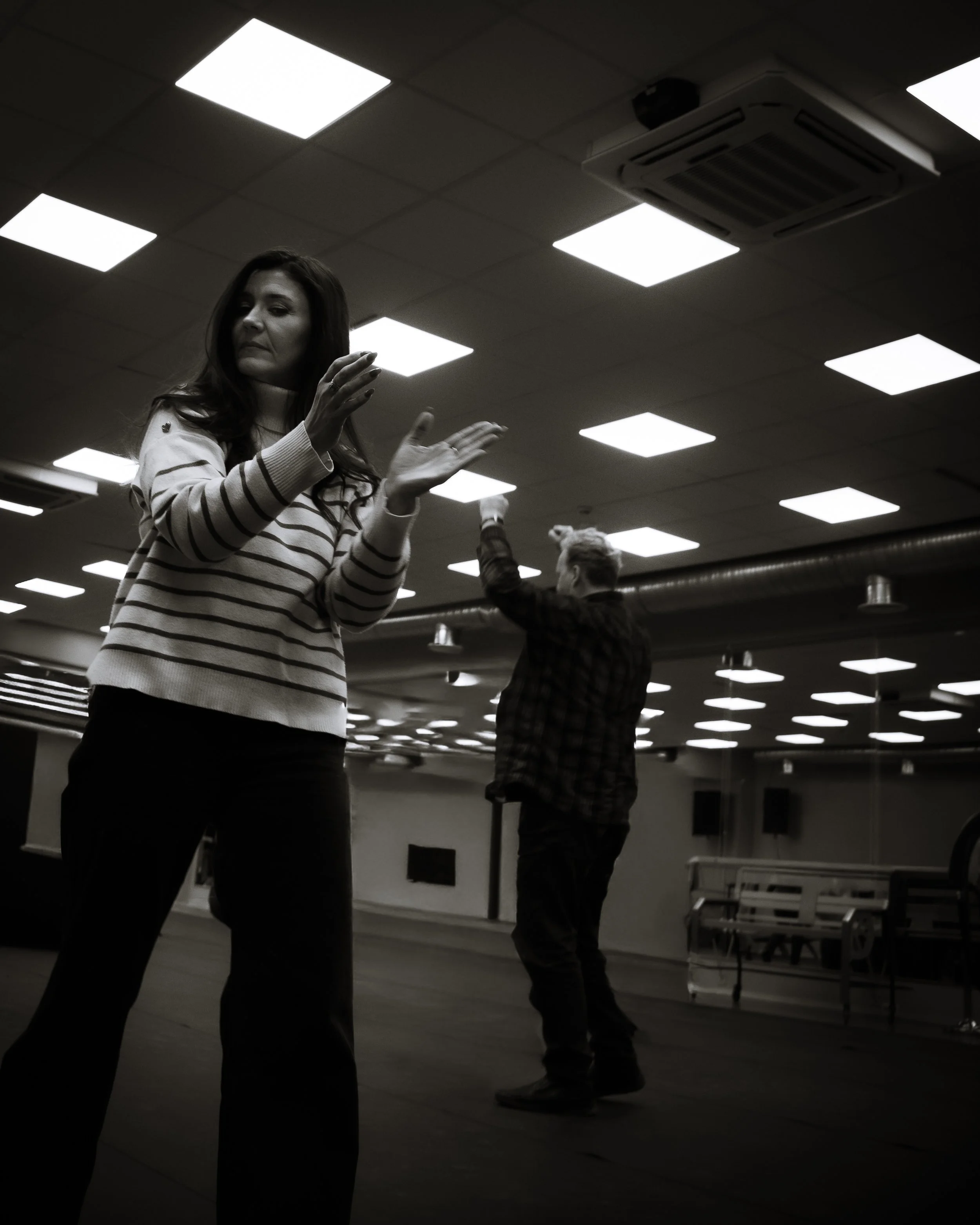 A woman in a striped sweater is dancing in a room with a man in a plaid shirt following her lead, under ceiling fluorescent lights.