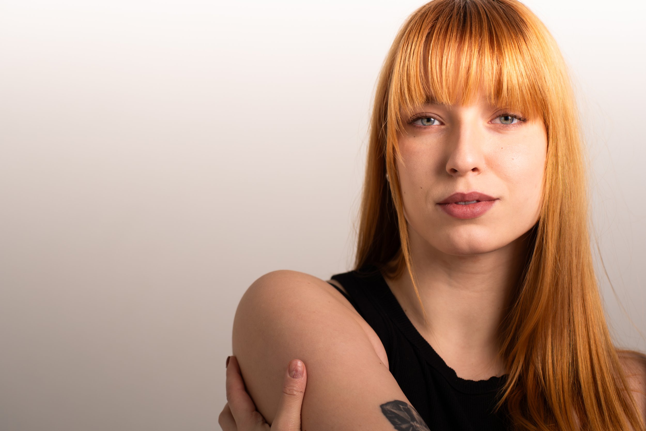 A woman with long red hair and blue eyes is smiling slightly, looking directly at the camera. She is wearing a black sleeveless top and has a tattoo on her left arm. She is standing against a plain, light-colored background.