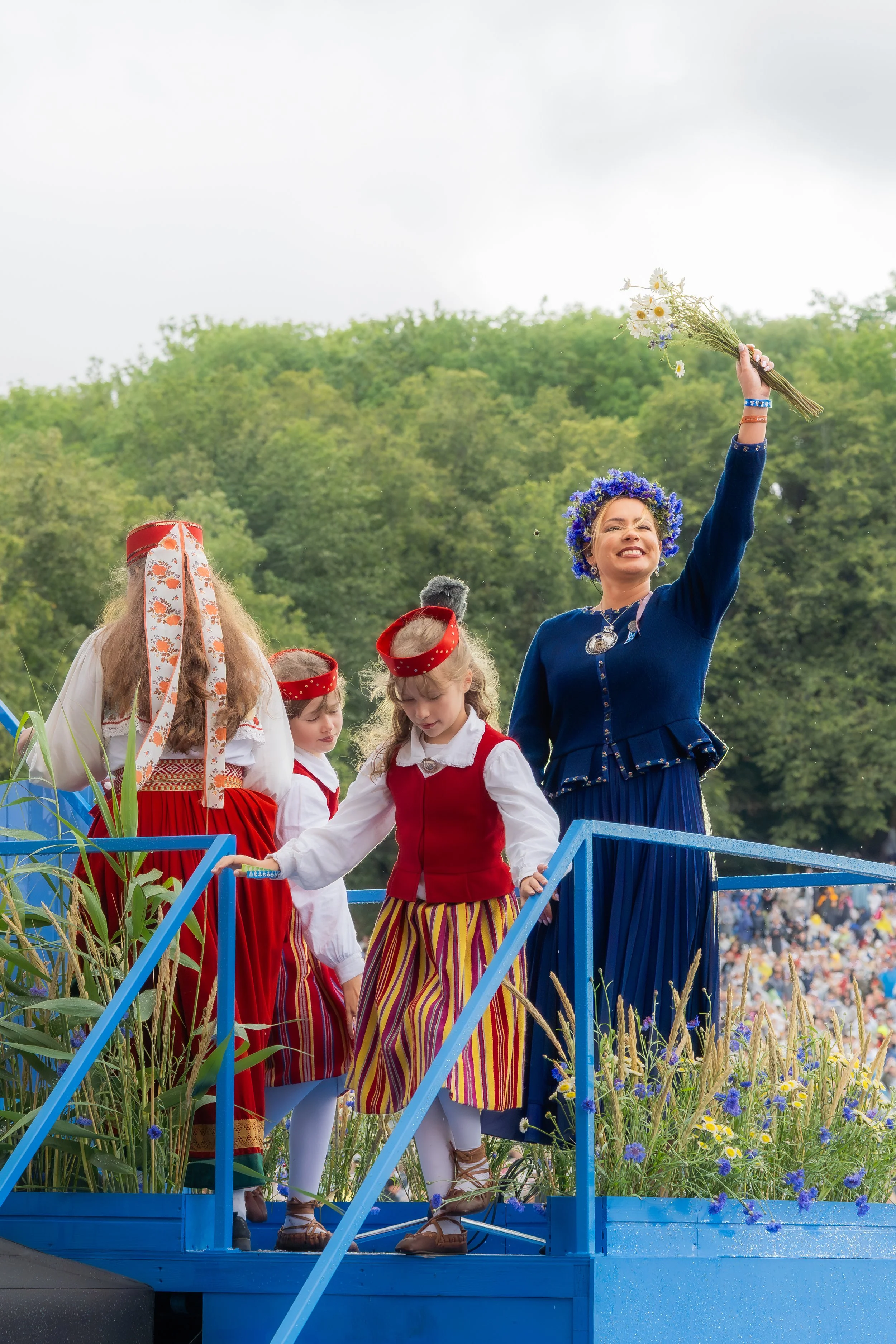 People dressed in traditional folk costumes celebrating on a decorated float during a parade, with one woman holding a bouquet of flowers and wearing a floral crown, and children in red and yellow costumes. Green trees and a crowd are visible in the 