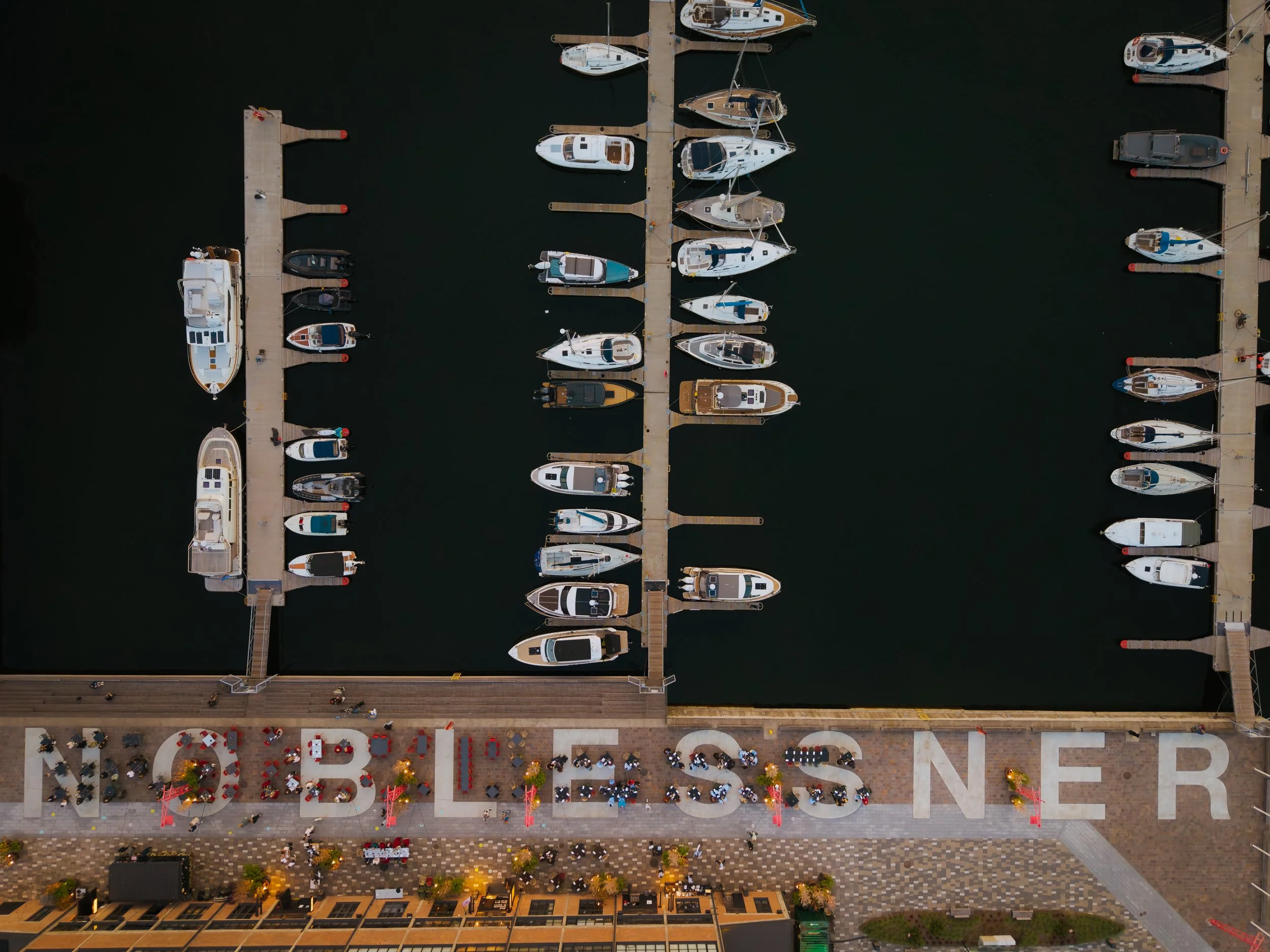 An aerial view of a marina with boats docked along piers, and an outdoor dining area with tables and umbrellas on the waterfront promenade.