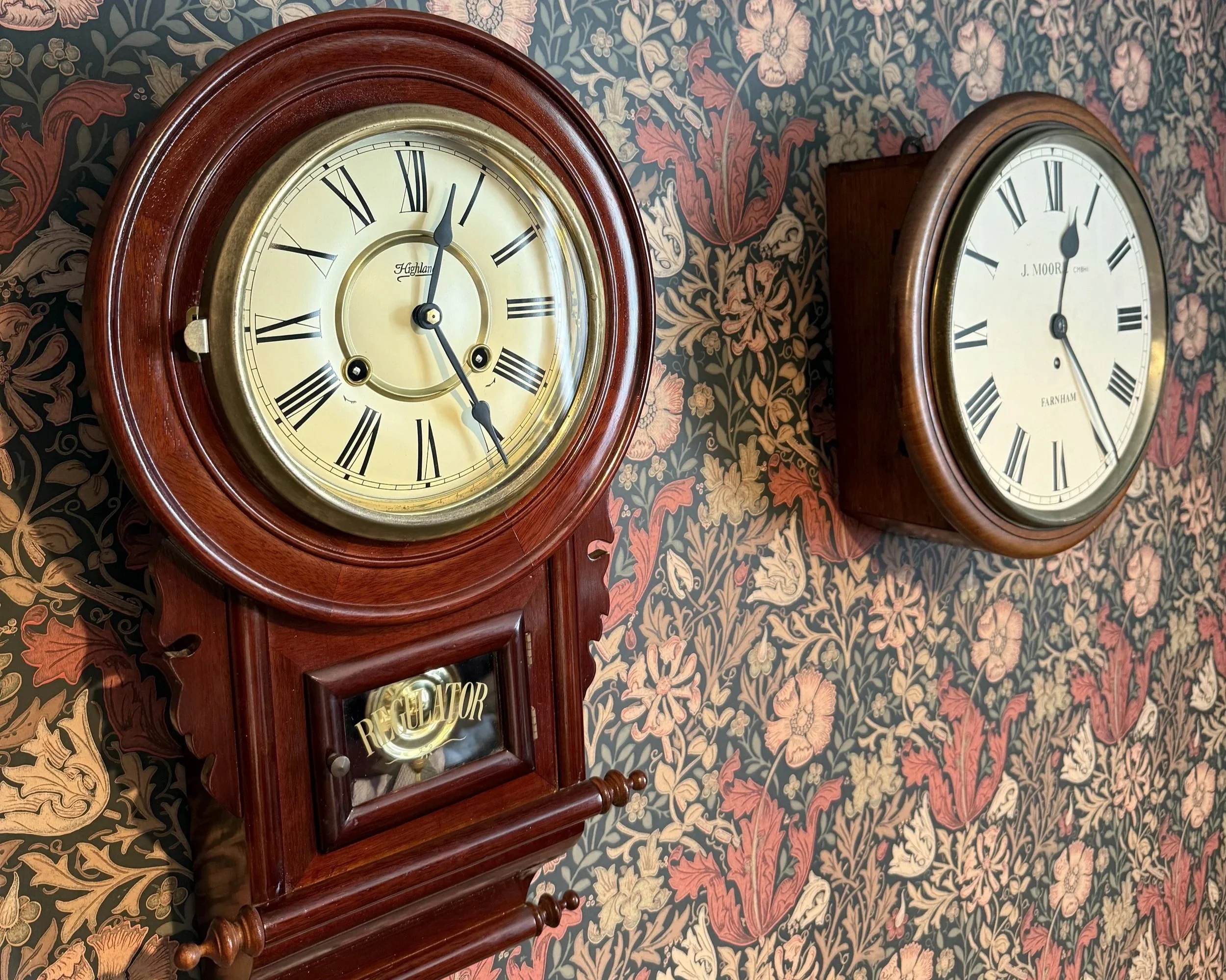 Close-up of a vintage wooden clock with a pale yellow face and black Roman numerals, showing the time as 12:07, mounted on a wall with floral patterned wallpaper.