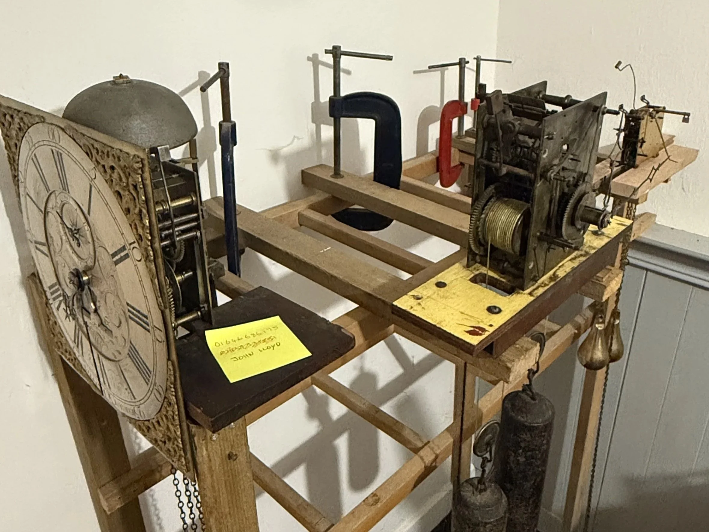 A wooden clockwork mechanism with gears and chains mounted on a wooden frame, with a decorative clock face on the left and various metal rods and parts connected to it.