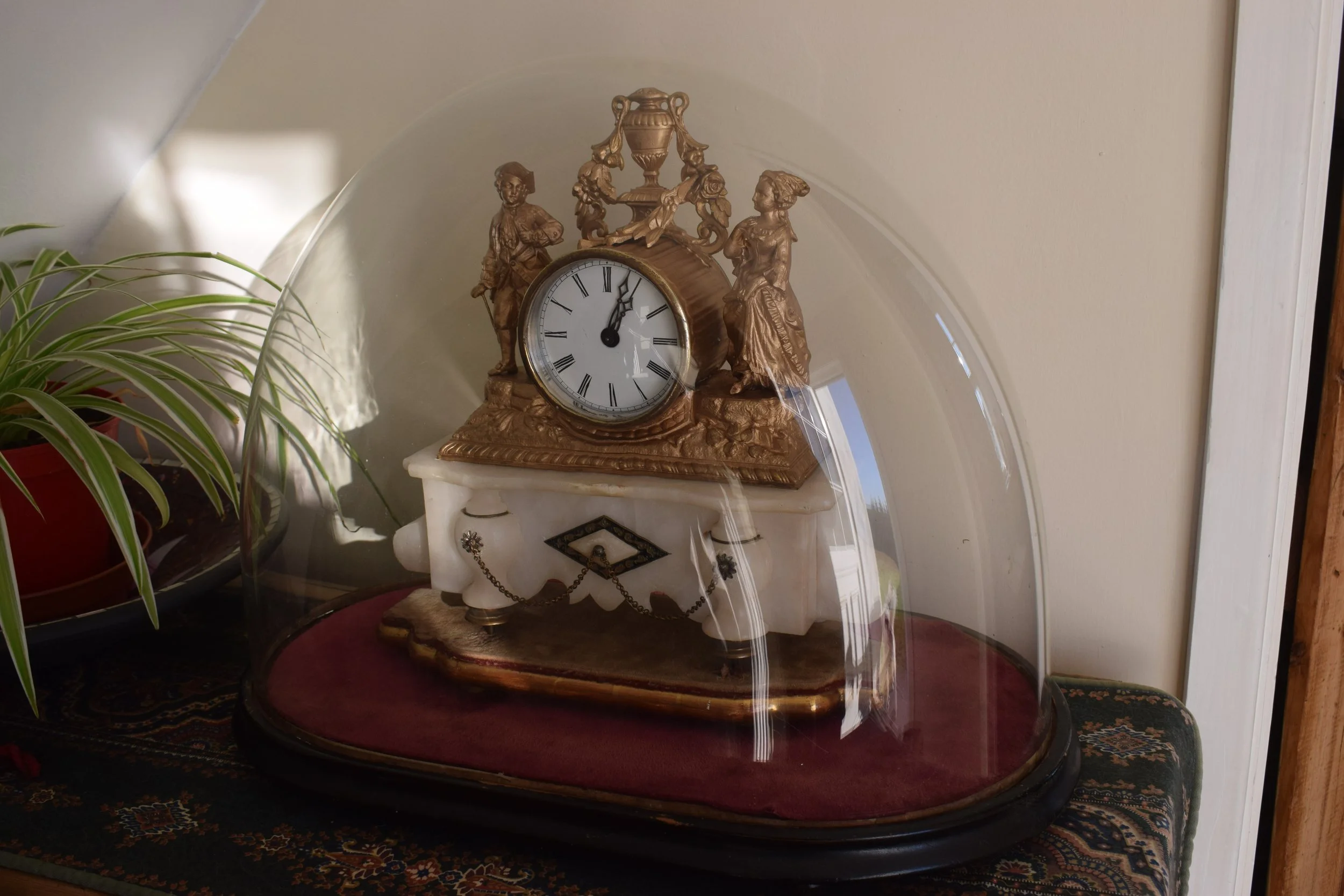 An ornate antique mantle clock with a gold finish, featuring figurines of a man and woman, and decorative urn on top, placed on a marble stand. The clock is situated on a round table with a dark surface, against a plain wall, with a plant with long, narrow leaves nearby.