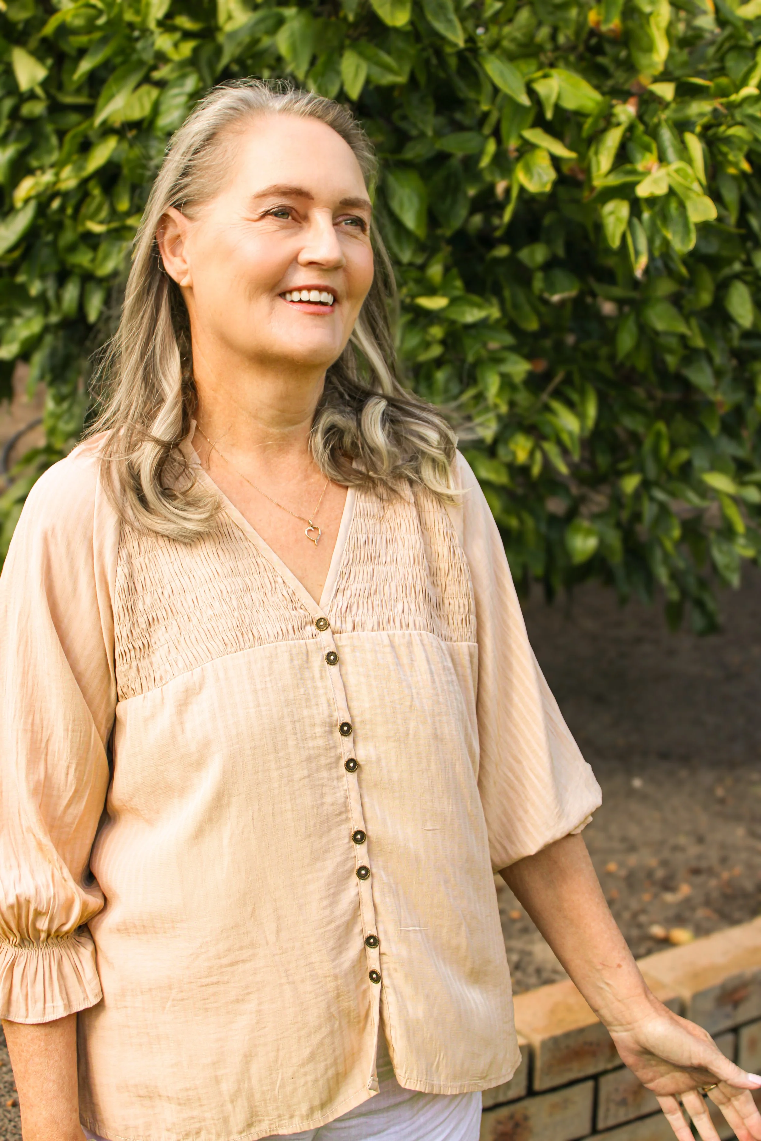 A woman with long gray hair smiling outdoors in front of green foliage, wearing a beige blouse and a necklace with a heart pendant.