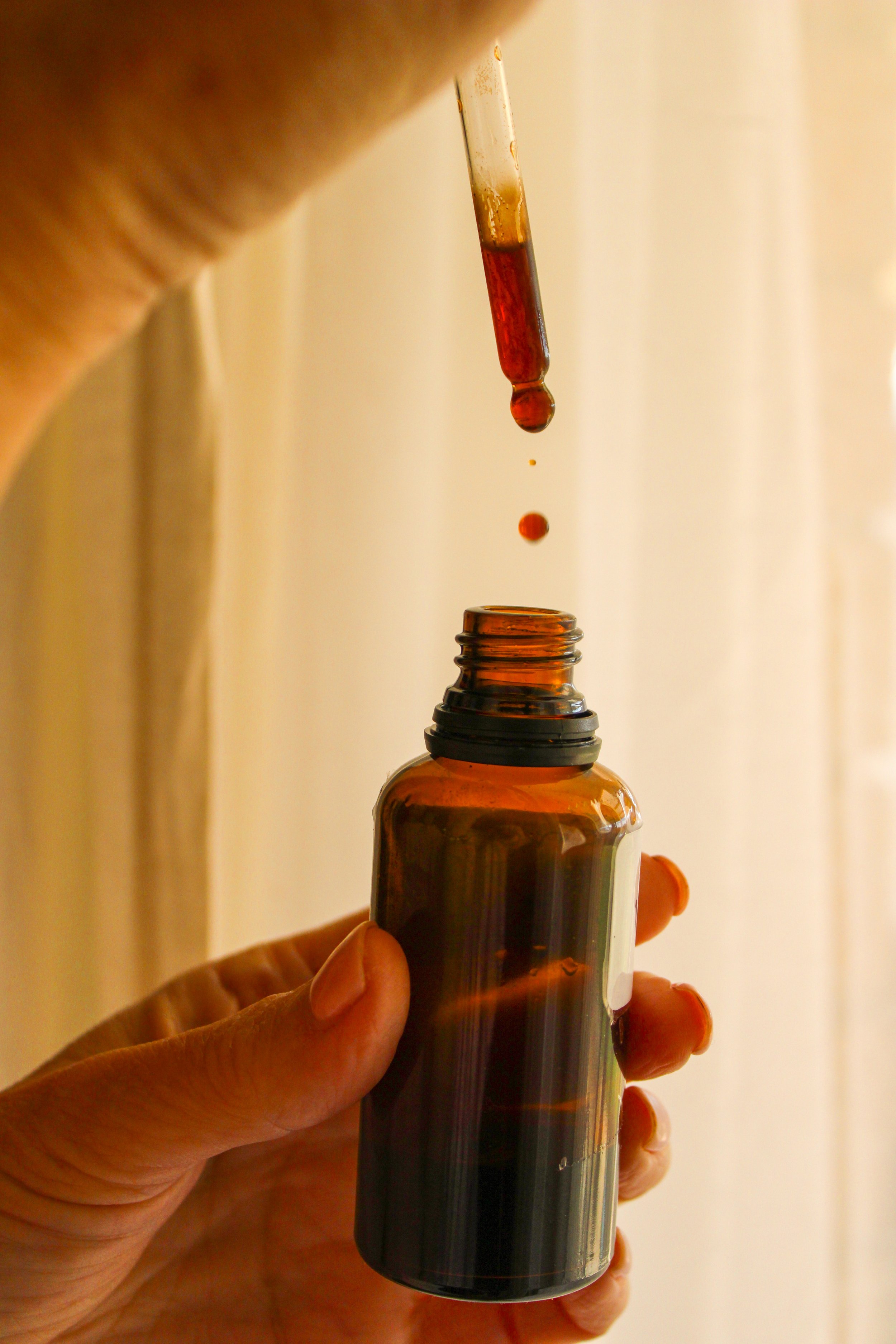 A person holding an amber glass dropper bottle with some liquid inside, and the dropper cap with a drop of liquid about to fall into the bottle.