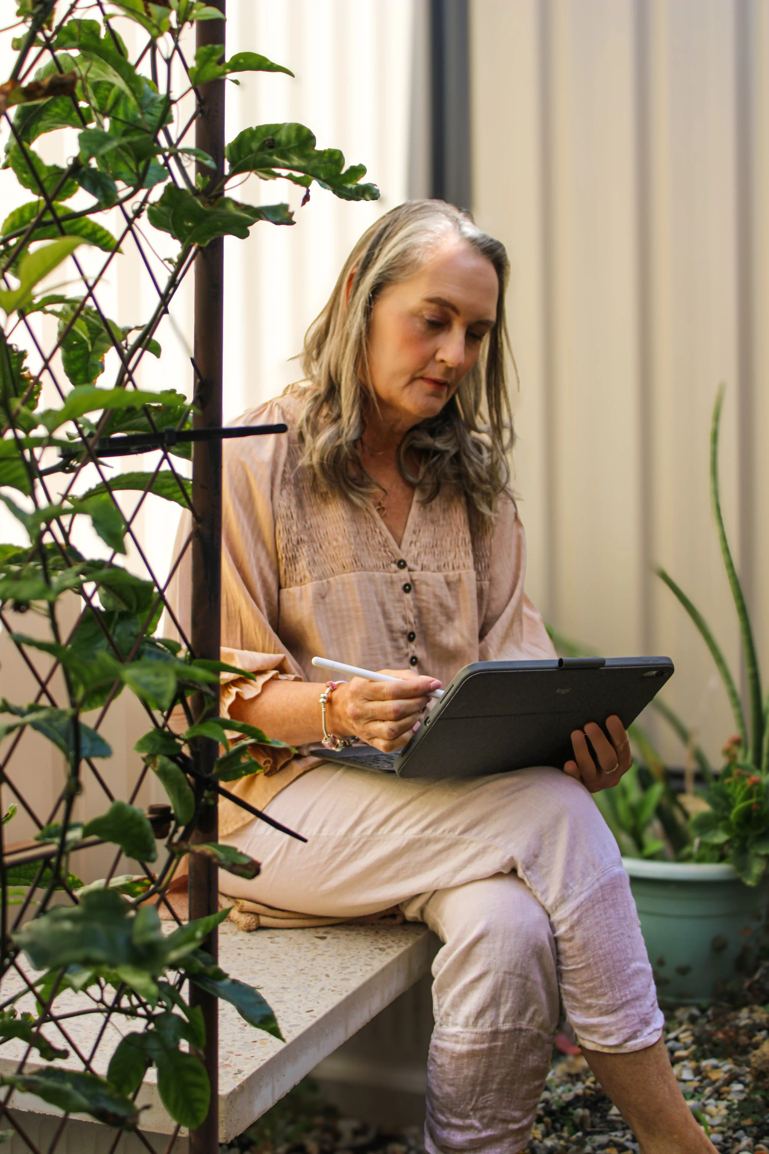 A woman with blonde hair sitting on a concrete bench in a garden, using a tablet device. She is wearing beige clothing and surrounded by plants and greenery.