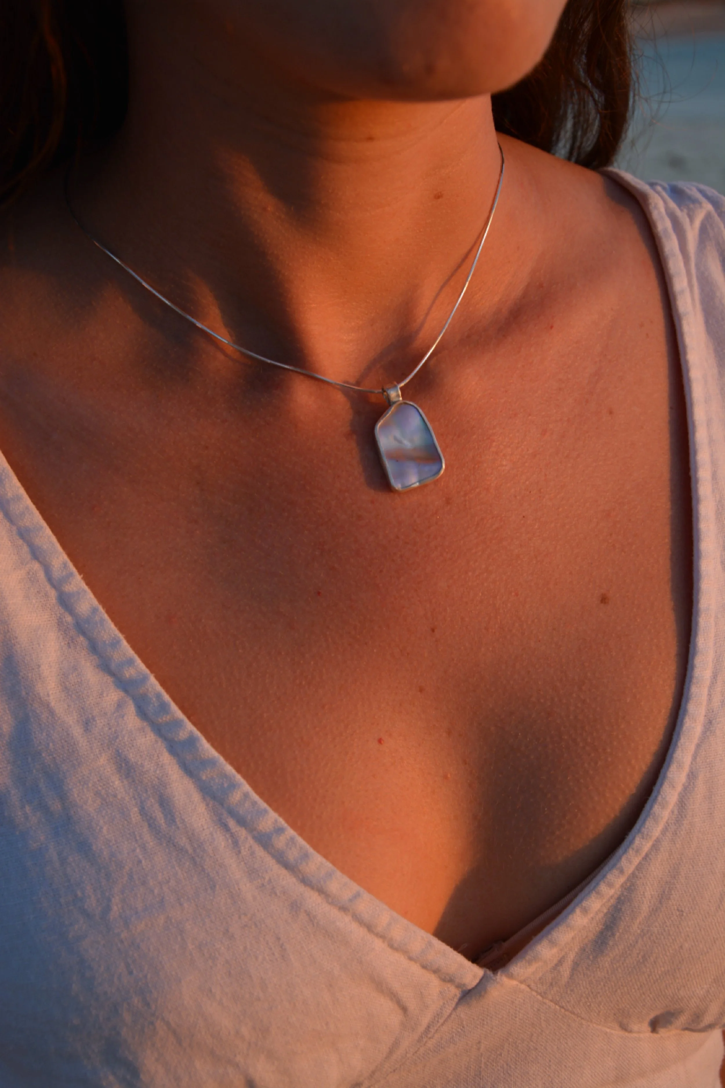Close-up of a woman wearing a silver necklace with a large iridescent abalone pendant, captured during golden hour.