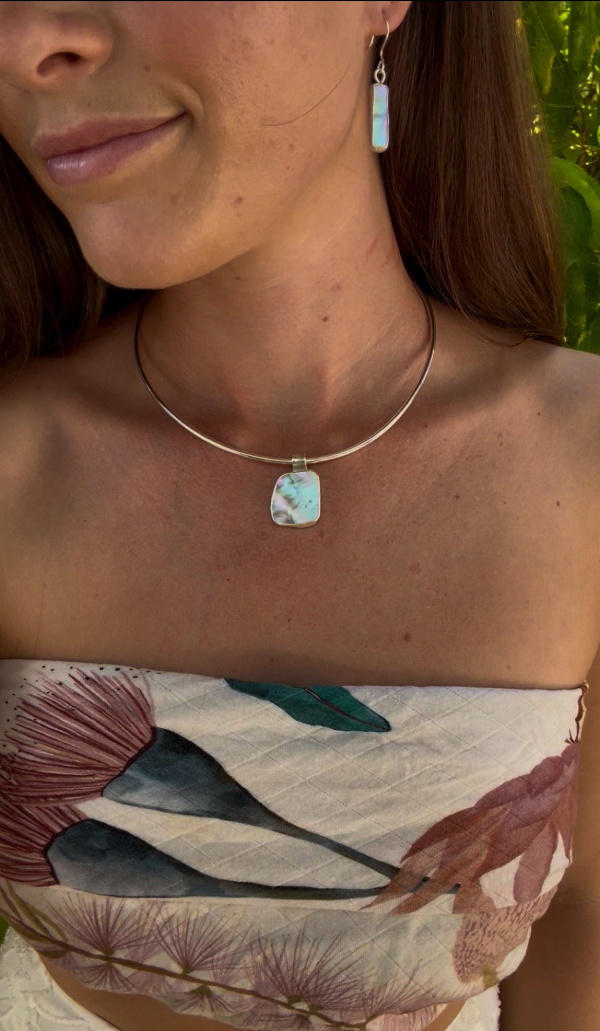 Close-up of a woman wearing a silver necklace and matching earrings with iridescent stones, showing her smiling lips, shoulder, and part of her patterned dress with floral designs.