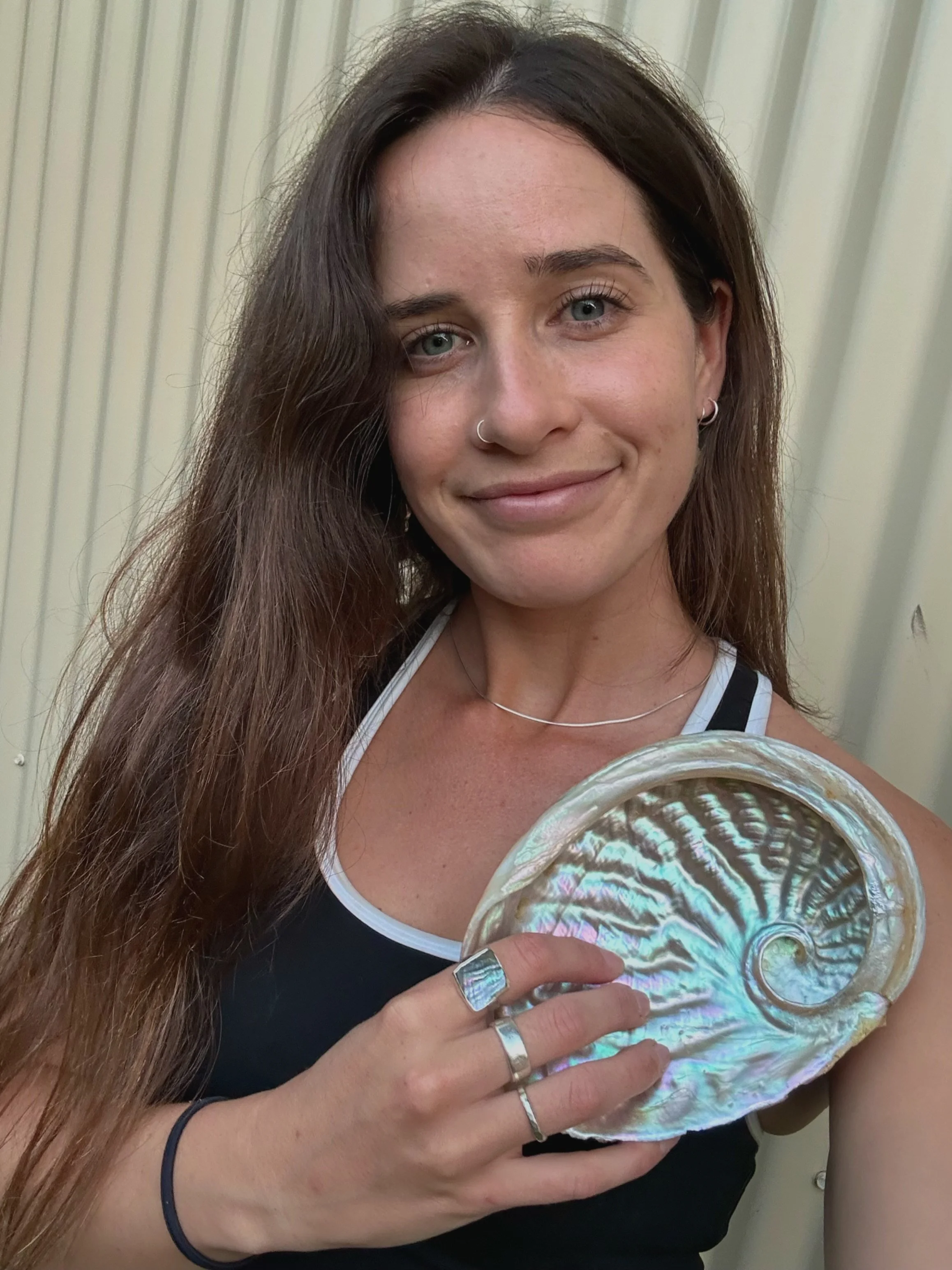 A woman with long brown hair and blue eyes is smiling at the camera while holding a large, shiny seashell in her hand. She is wearing a black sleeveless top, a silver ring on her index finger, a silver bracelet, and small hoop earrings. The background features vertical beige or light-colored blinds.