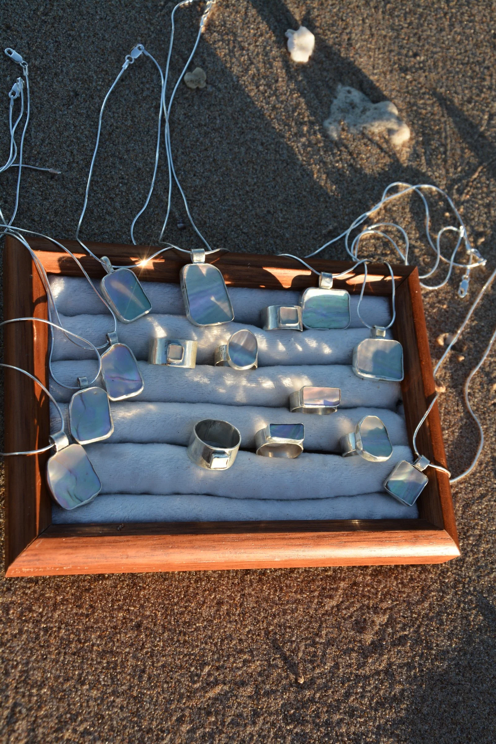 A wooden jewelry box containing abalone silver jewellery on a sandy surface with seashells nearby.