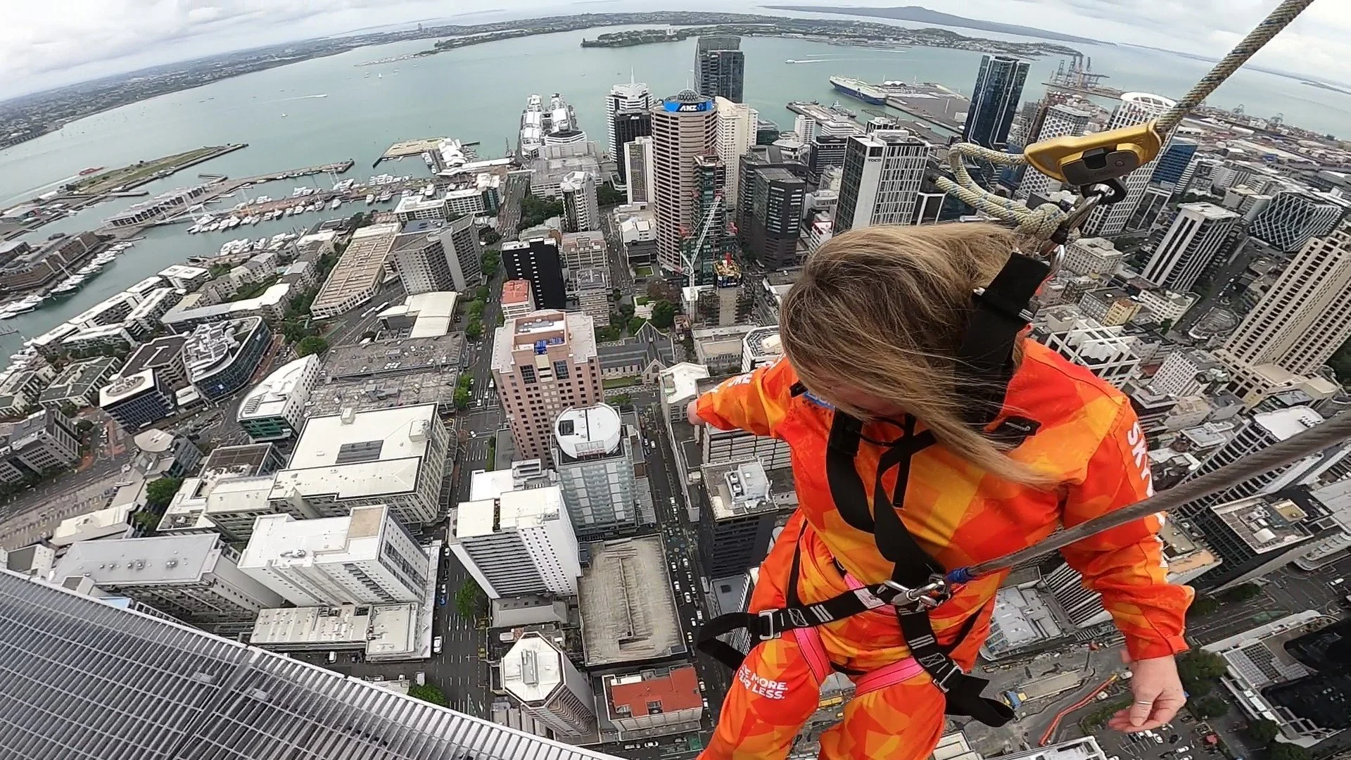 A woman wearing bright orange safety gear leans over the edge of a skyscraper, overlooking a cityscape with tall buildings, a harbor, and water extending into the distance