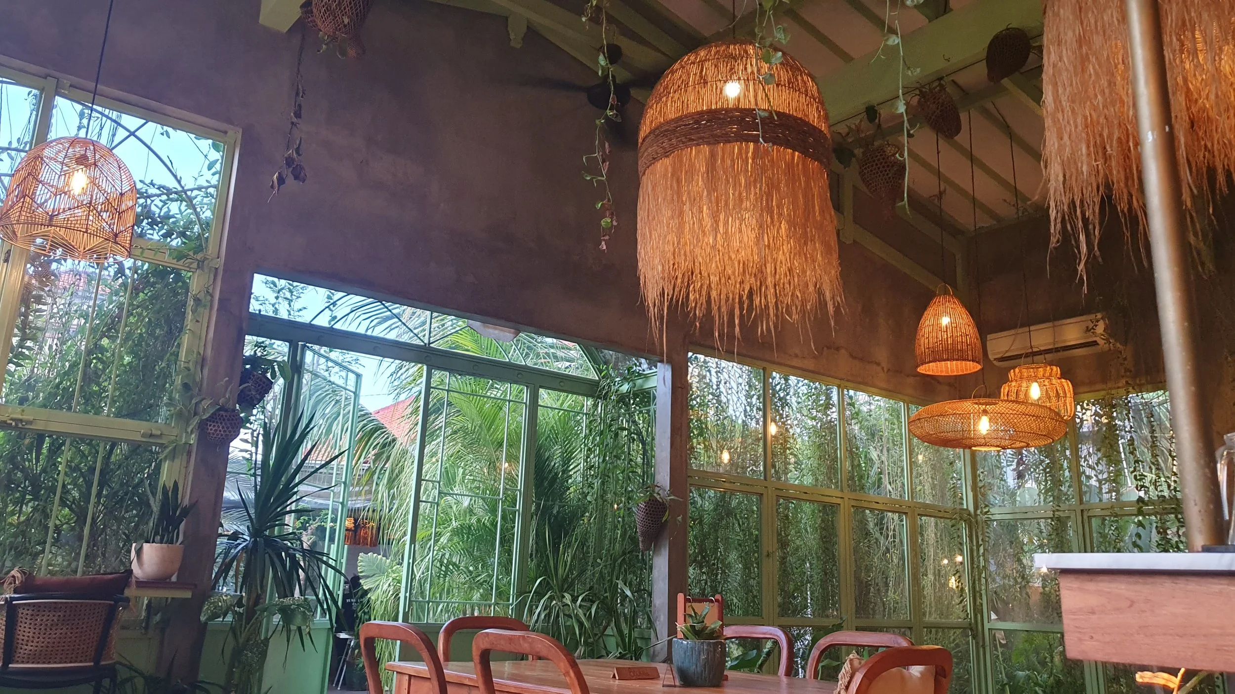 Interior of a cozy cafe or restaurant with large windows, hanging wicker lamps, and lush green plants inside.