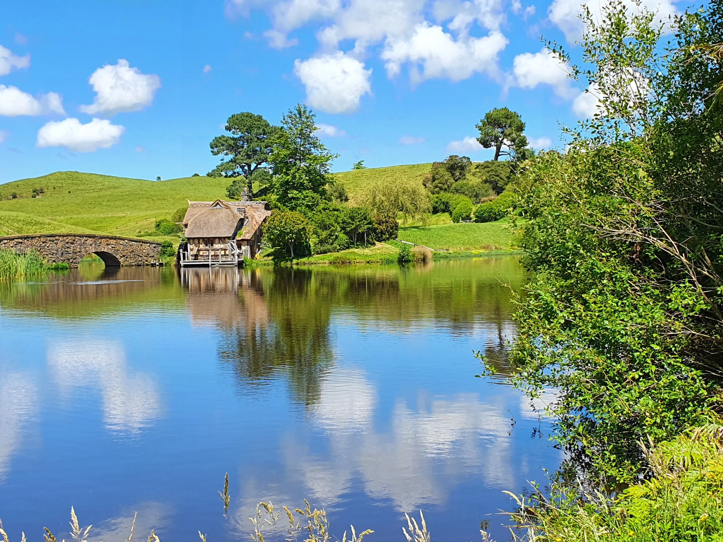 A peaceful landscape featuring a calm river reflecting the blue sky and white clouds, surrounded by lush green trees and grassy hills with a small stone bridge and a rustic water mill.