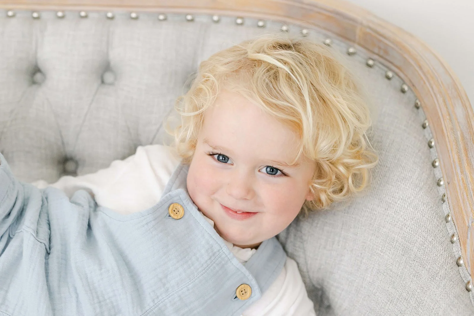 A young boy with curly blonde hair and blue eyes, lying on a cushioned, gray upholstered armchair with a wooden frame and decorative nailhead trim.